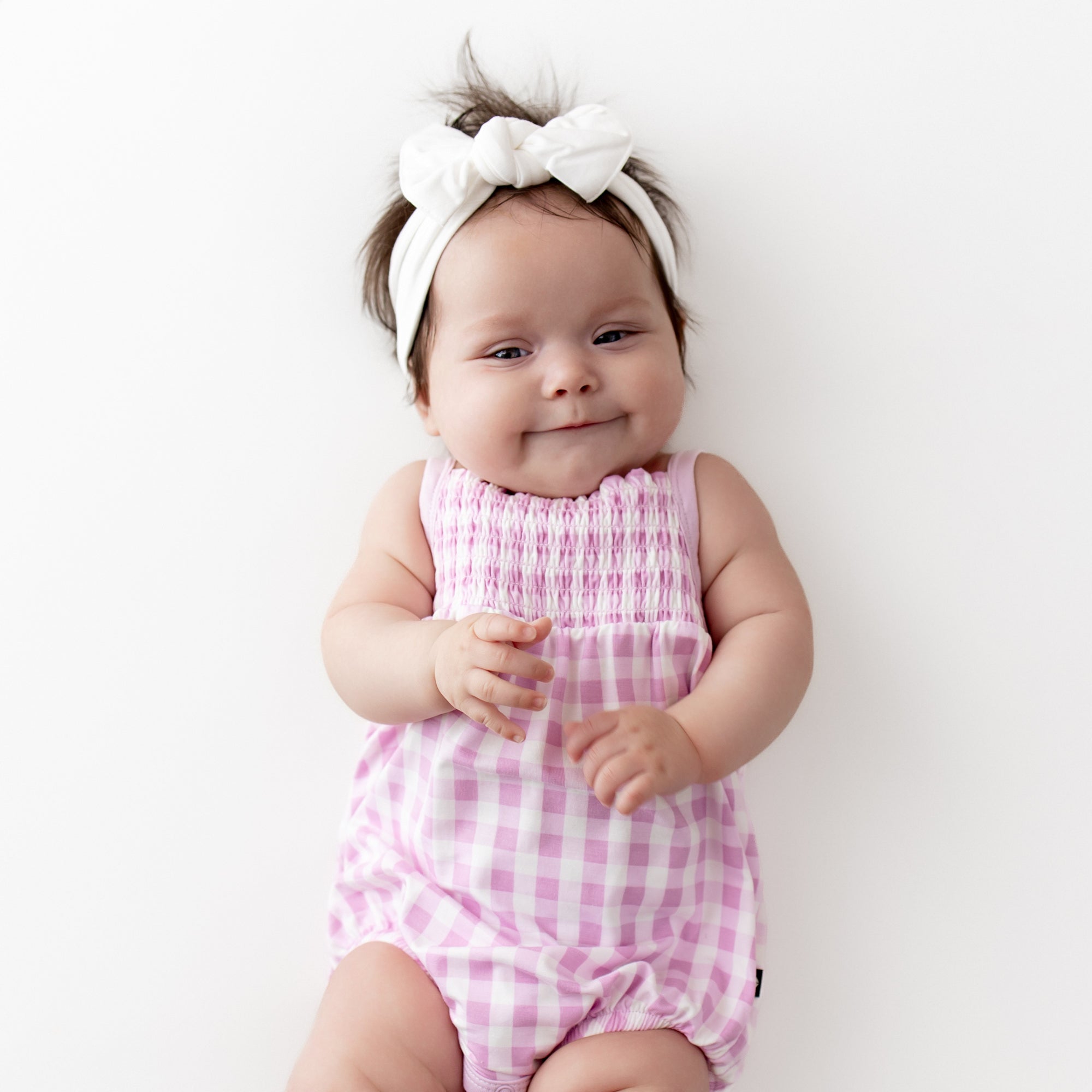 Close up of infant wearing the Smocked Bubble Romper in Gingham Thistle with a white knotted bow headband