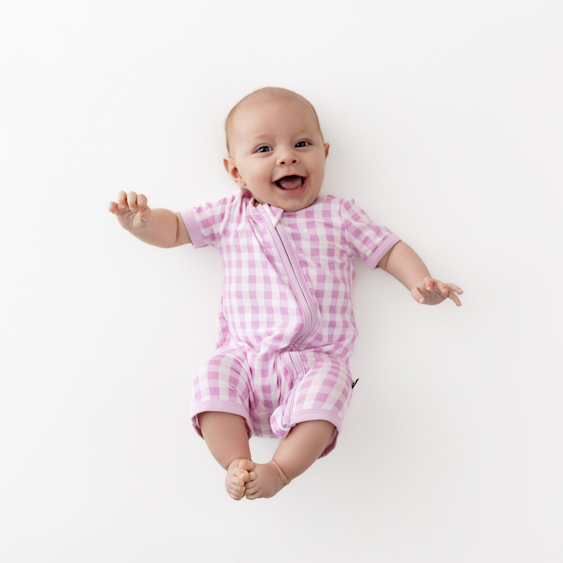 Smiling infant wearing the Zipper Shortall Romper in Gingham Thistle laying on a light cream surface