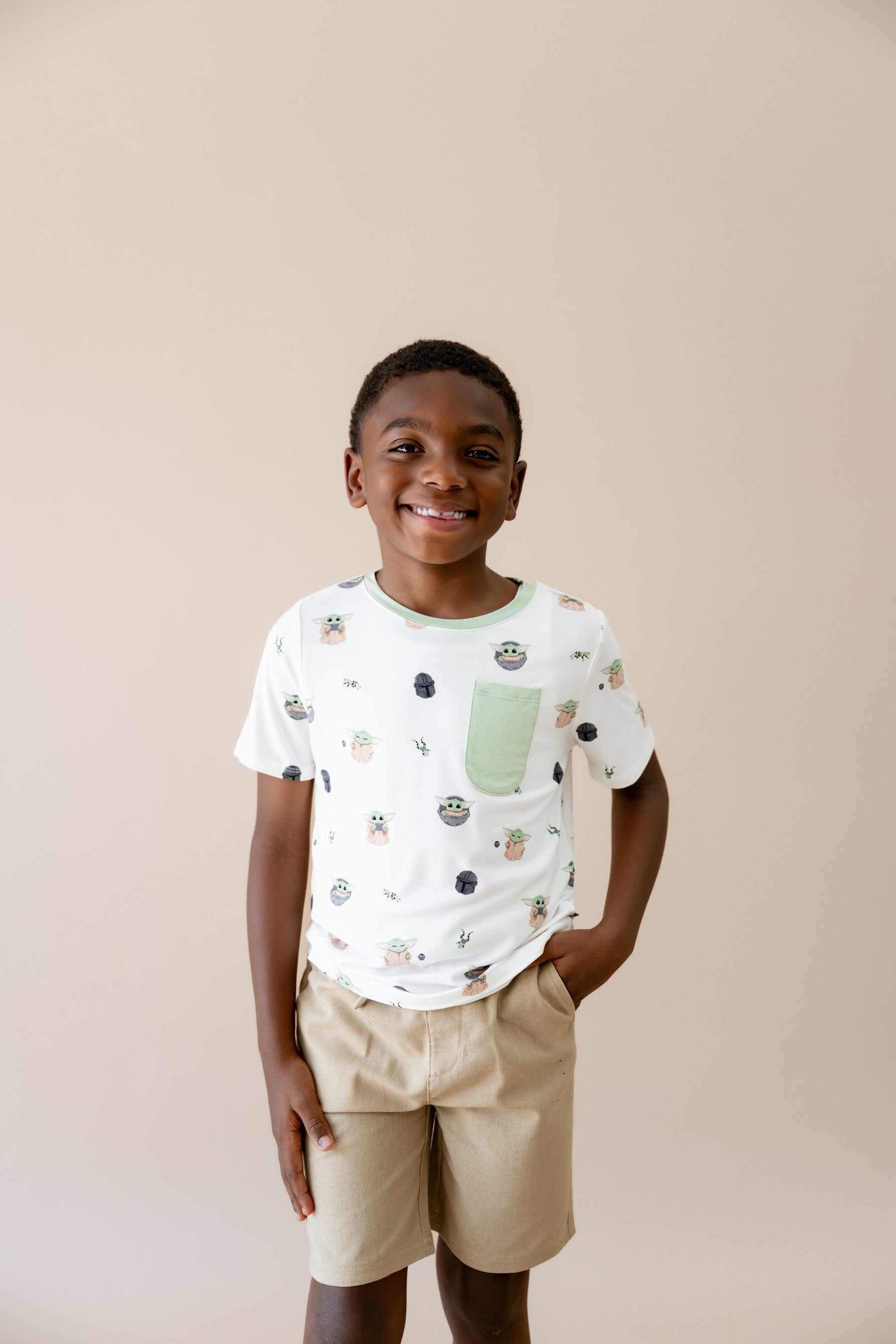 Smiling boy standing in front of a taupe background wearing the Toddler Crew Neck Tee in Grogu™ with tan shorts