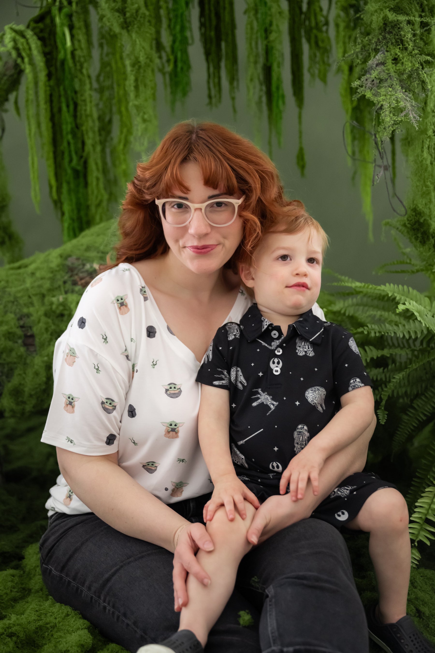 Young boy wearing the Toddler Short Sleeve Polo in STAR WARS™ sitting on his mother's lap who is wearing the Women's V-Neck in Grogu™