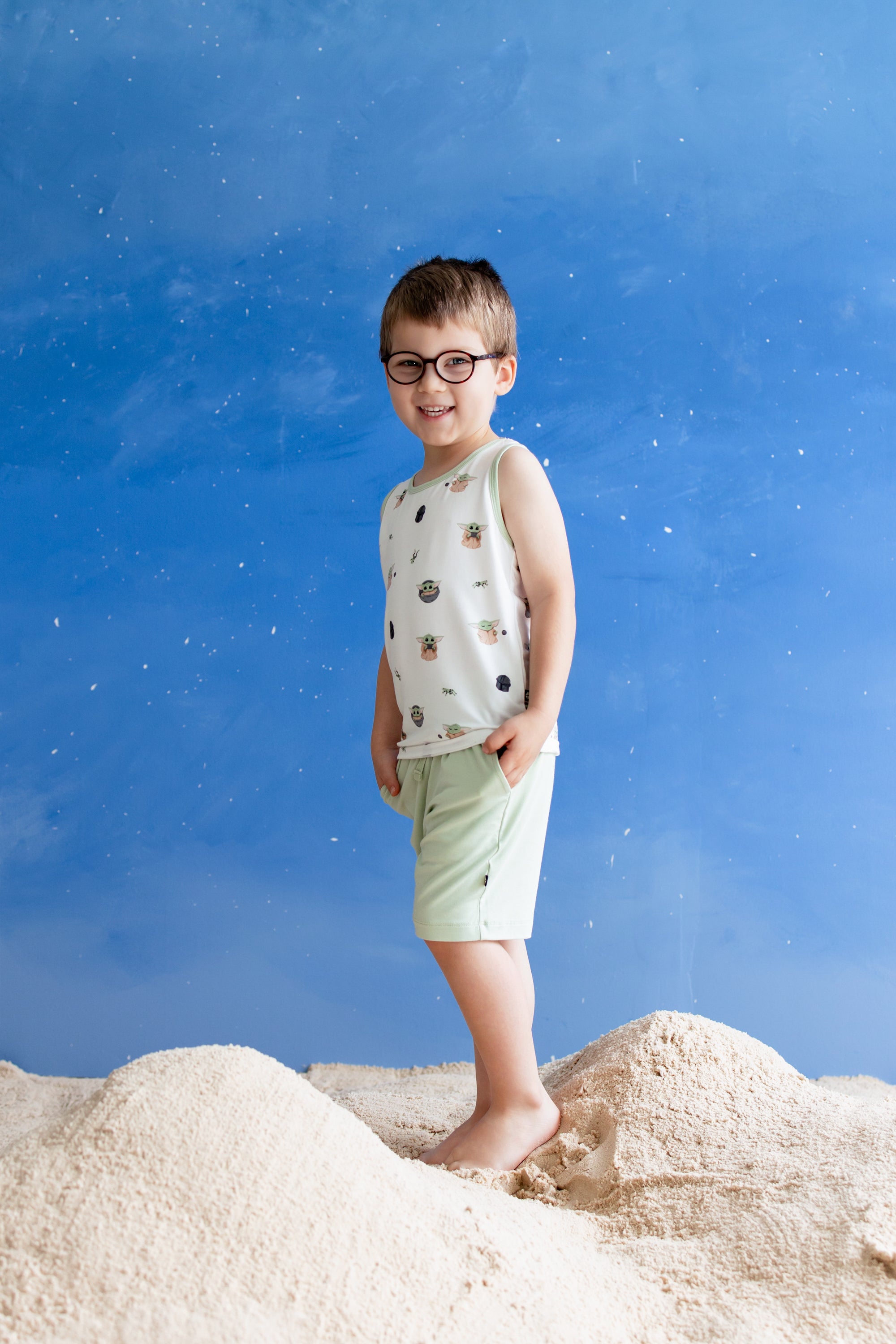 Young boy walking through sand in front of a starry blue background wearing the Toddler Tank Top in Grogu™ with light green shorts