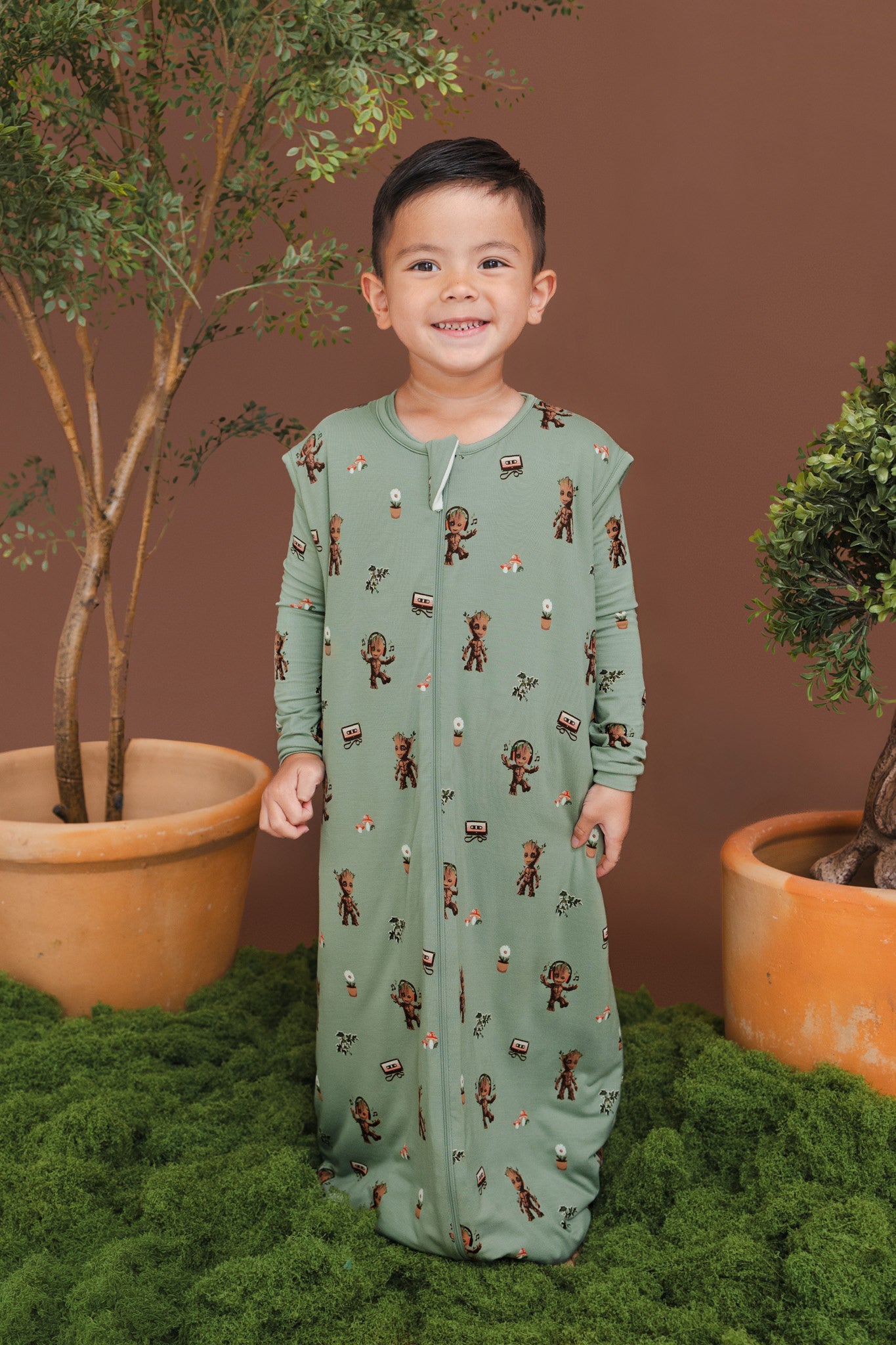 Boy smiling in Green Sleep Bag Walker in Groot 1.0 print standing between green plant pots and grass