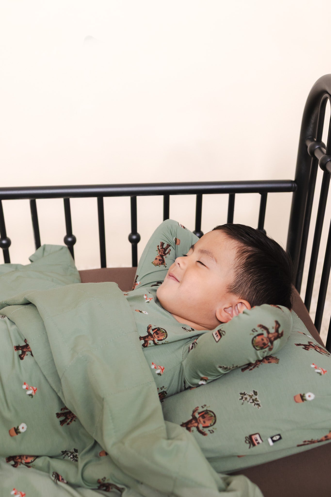 Boy lying on a bed under Green Toddler Blanket in Groot 1.0 print over a similar print pillow and wearing a similar print long sleeve pajama set