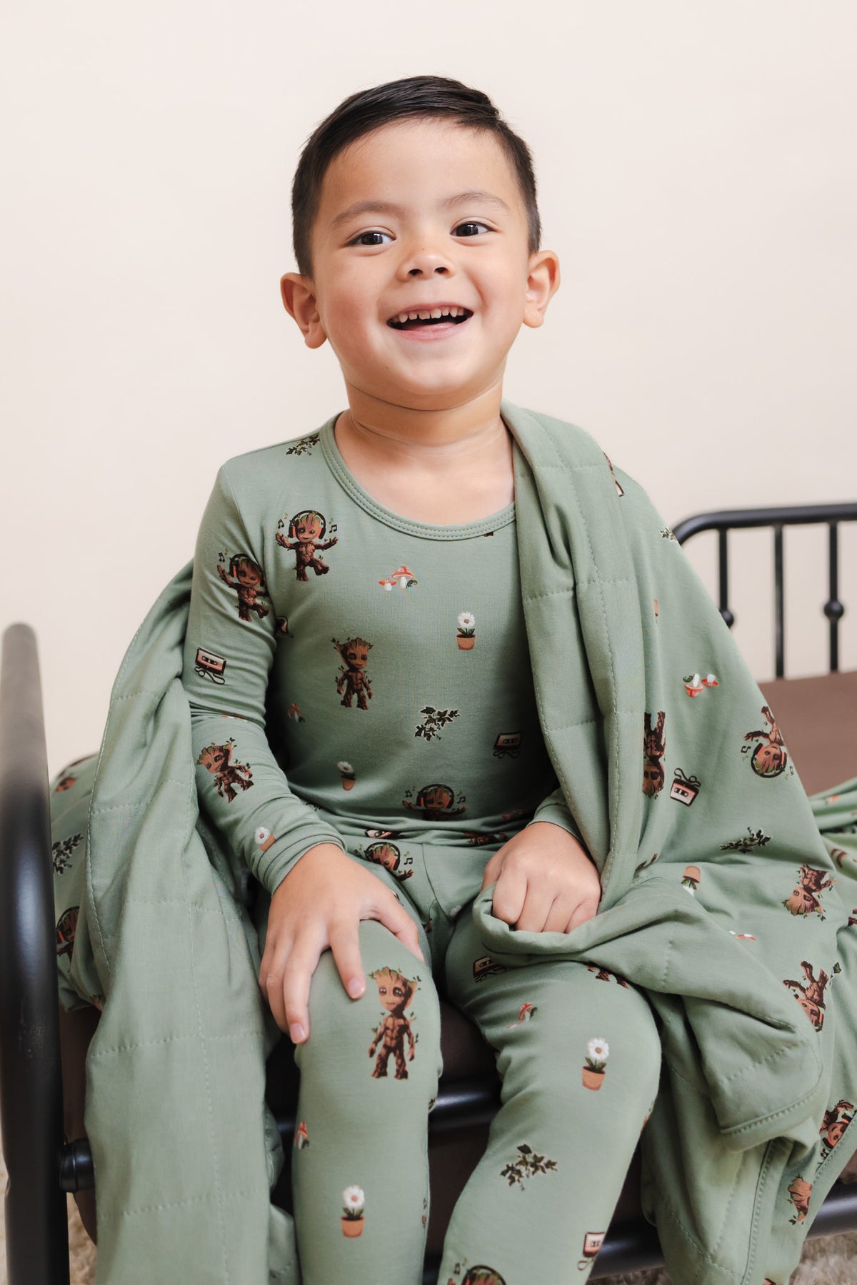Boy sitting with Green Toddler Blanket in Groot 1.0 print on a bed wearing a similar print long sleeve pajama set