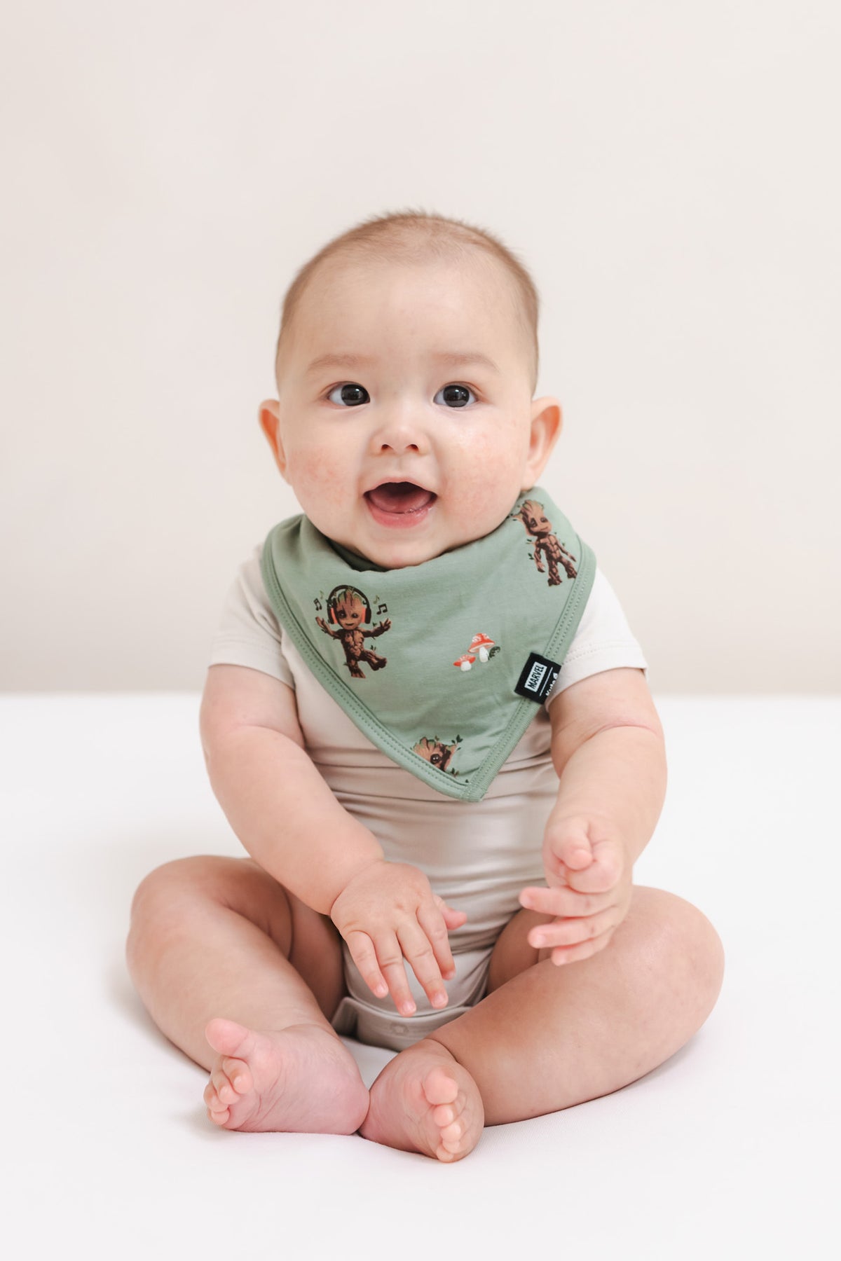 Baby sitting on a beige background wearing Green Bib in Groot print over a beige bodysuit
