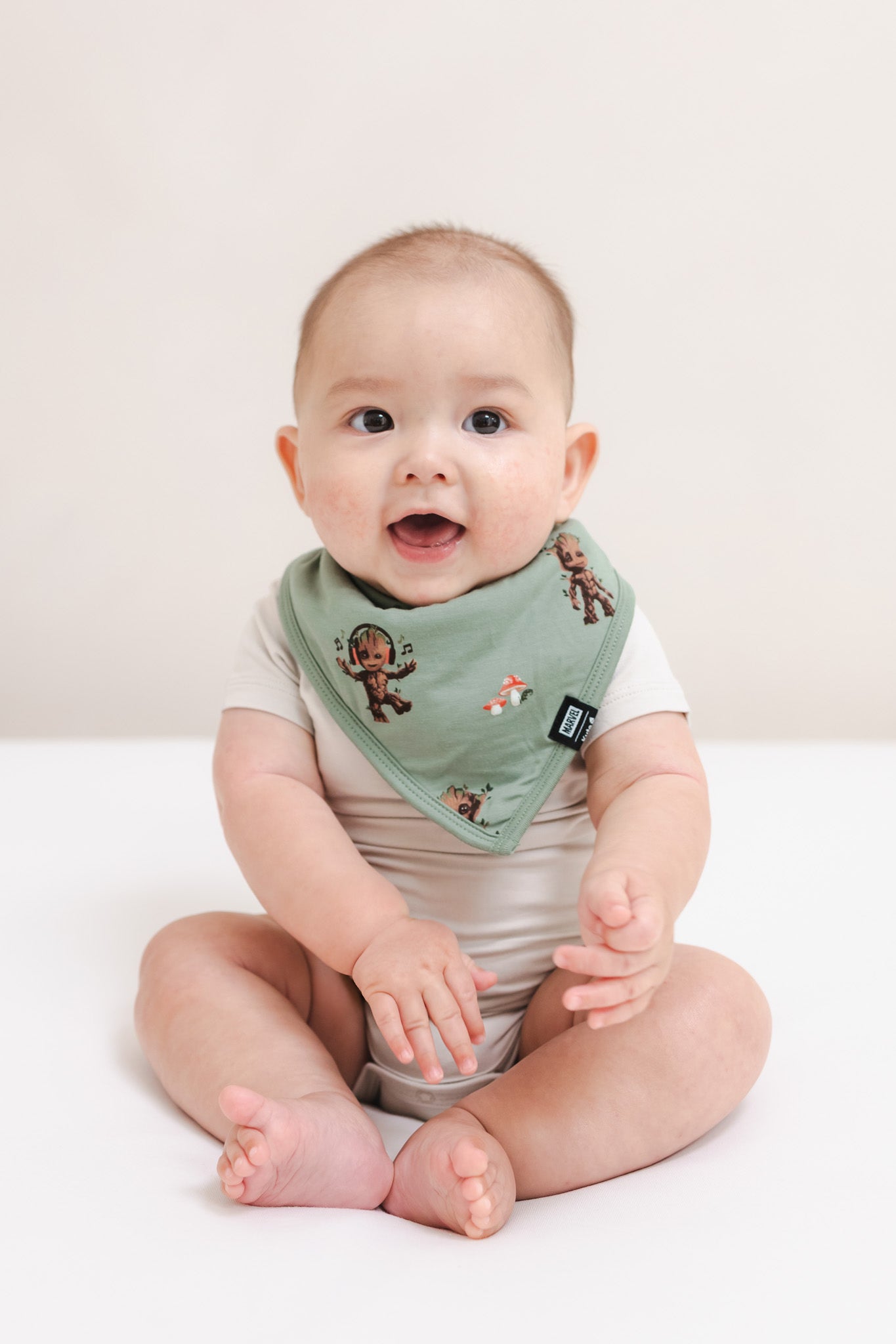 Baby sitting on a beige background wearing Green Bib in Groot print over a beige bodysuit