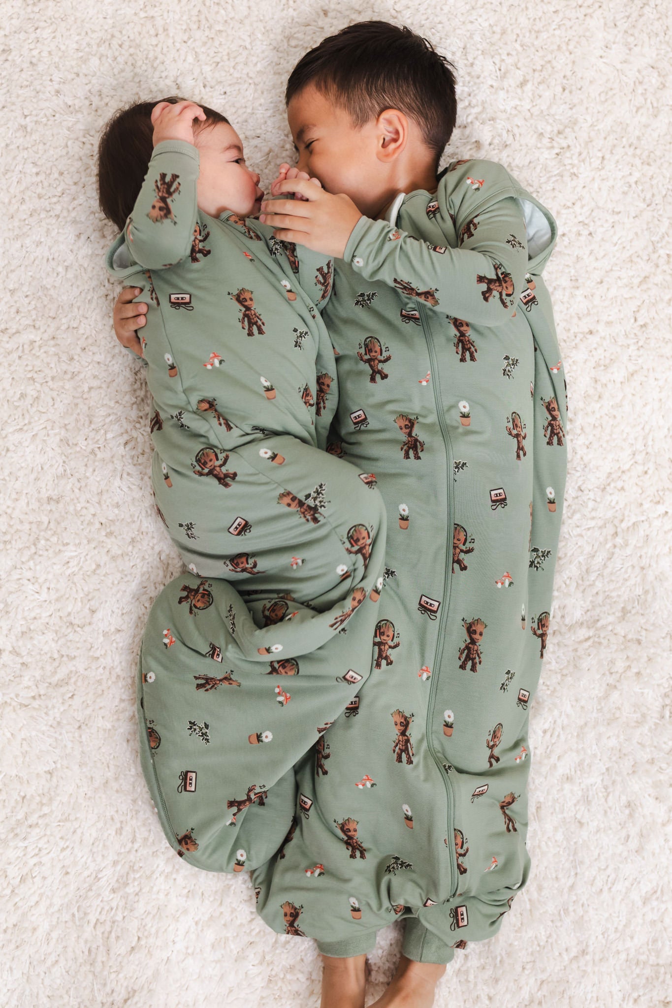 A Boy and a baby lying on a beige carpet cuddled together wearing Green Sleep Bag Walker in Groot 1.0 print and Sleep bag in Groot print
