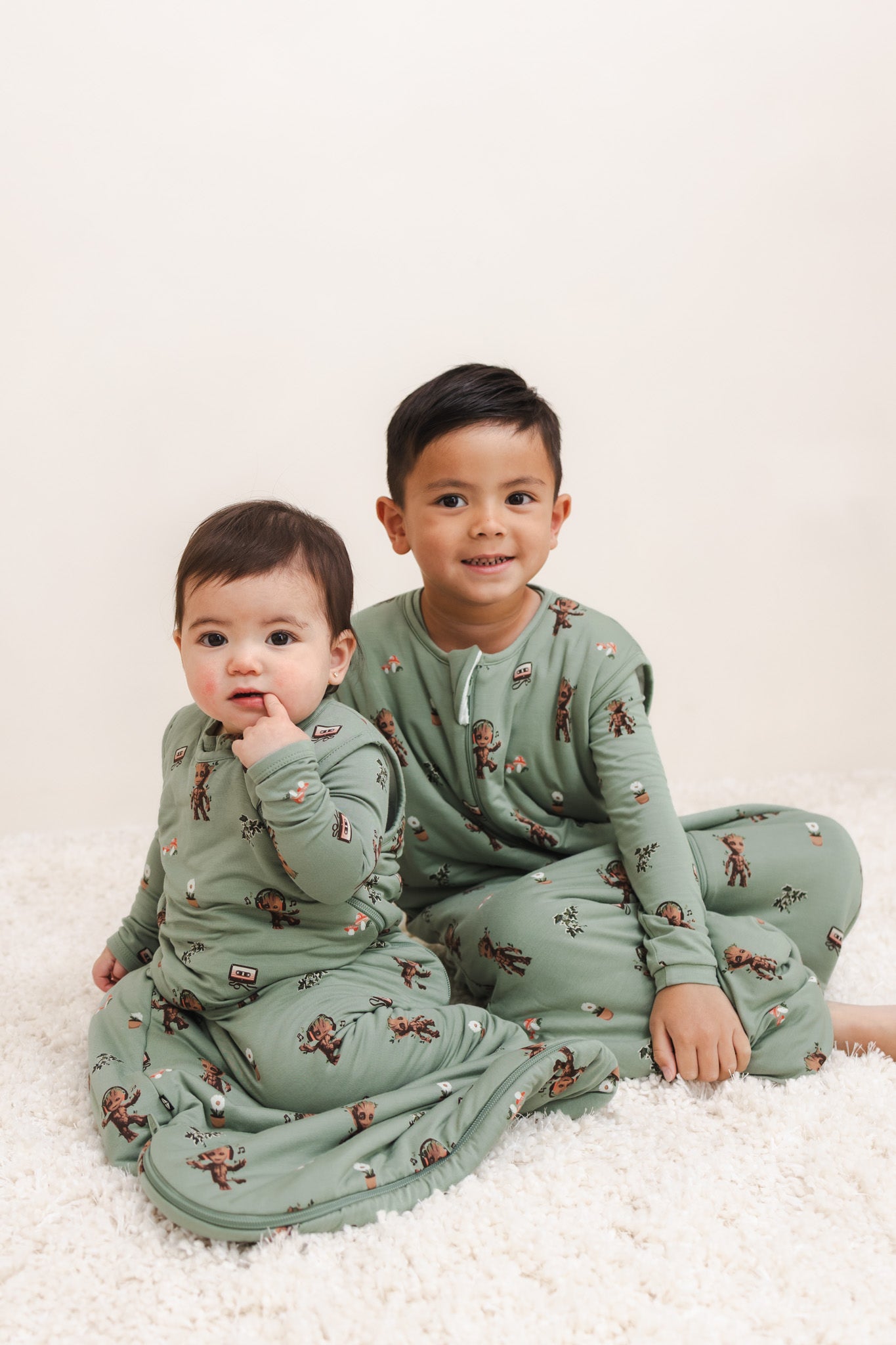 A Boy wearing Green Sleep Bag Walker in Groot 1.0 print sitting with a baby wearing Sleep bag in Groot print on a beige background