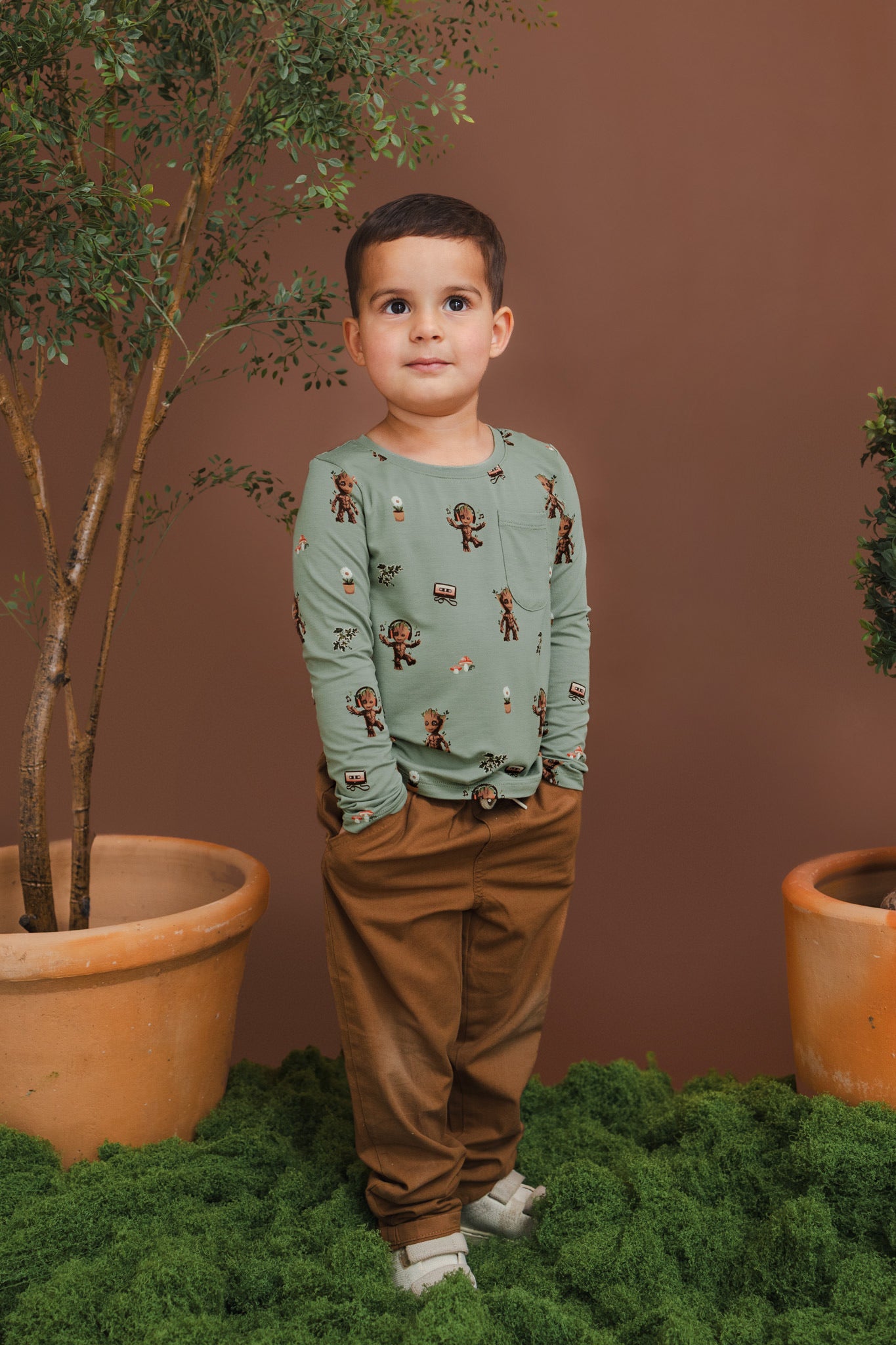 Boy posing in Green Long Sleeve Toddler Crew Neck Tee in Groot print over brown pants standing between plant pots and grass