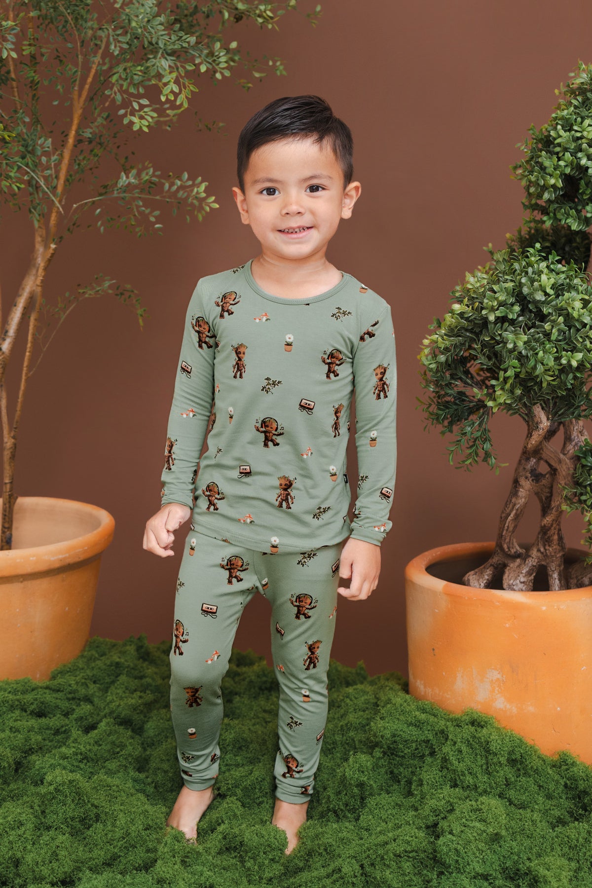 Boy standing on green grass and planted pots wearing Green Long Sleeve Pajamas in Groot print
