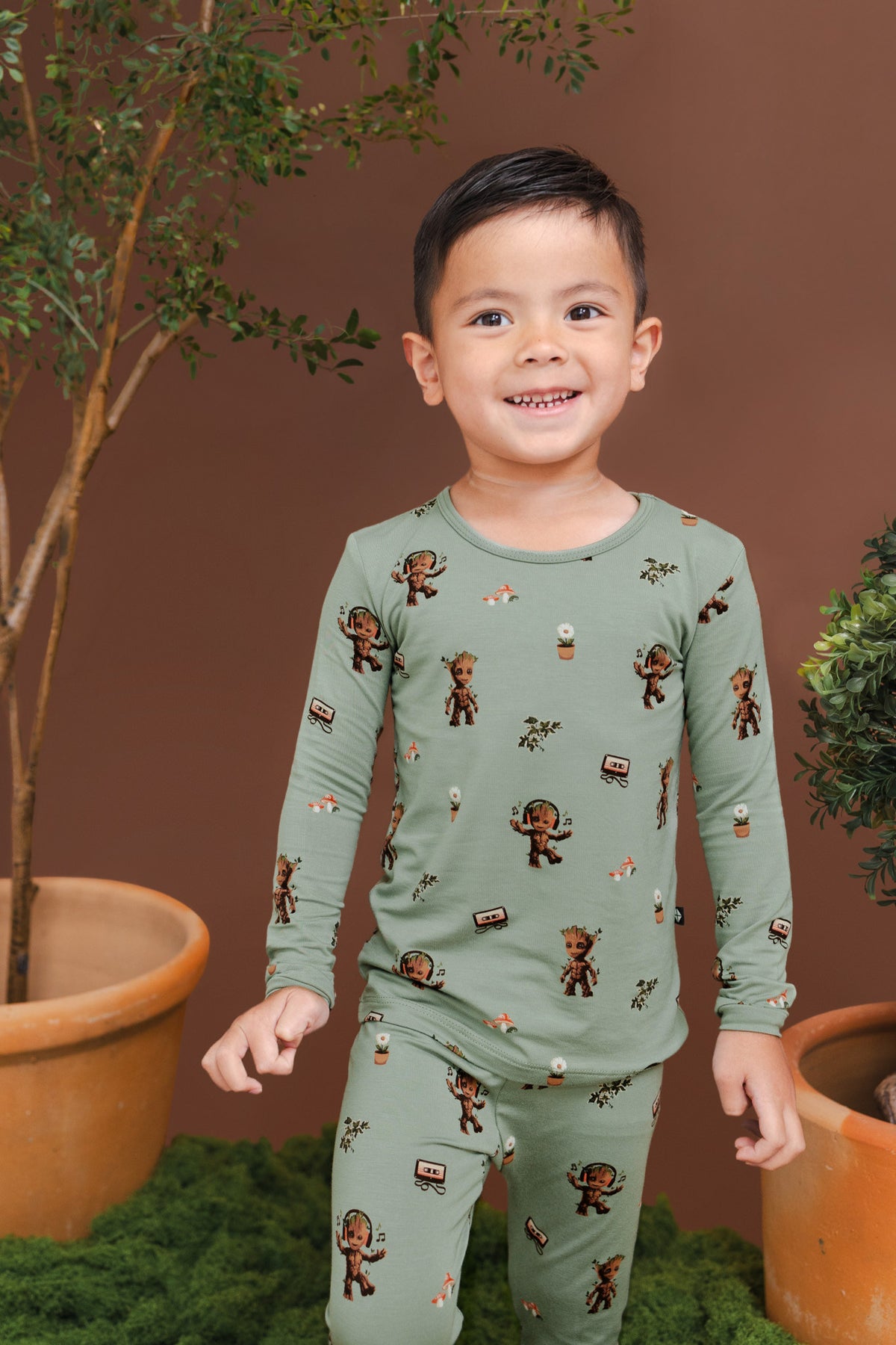 Boy smiling between planted pots wearing Green Long Sleeve Pajamas in Groot print