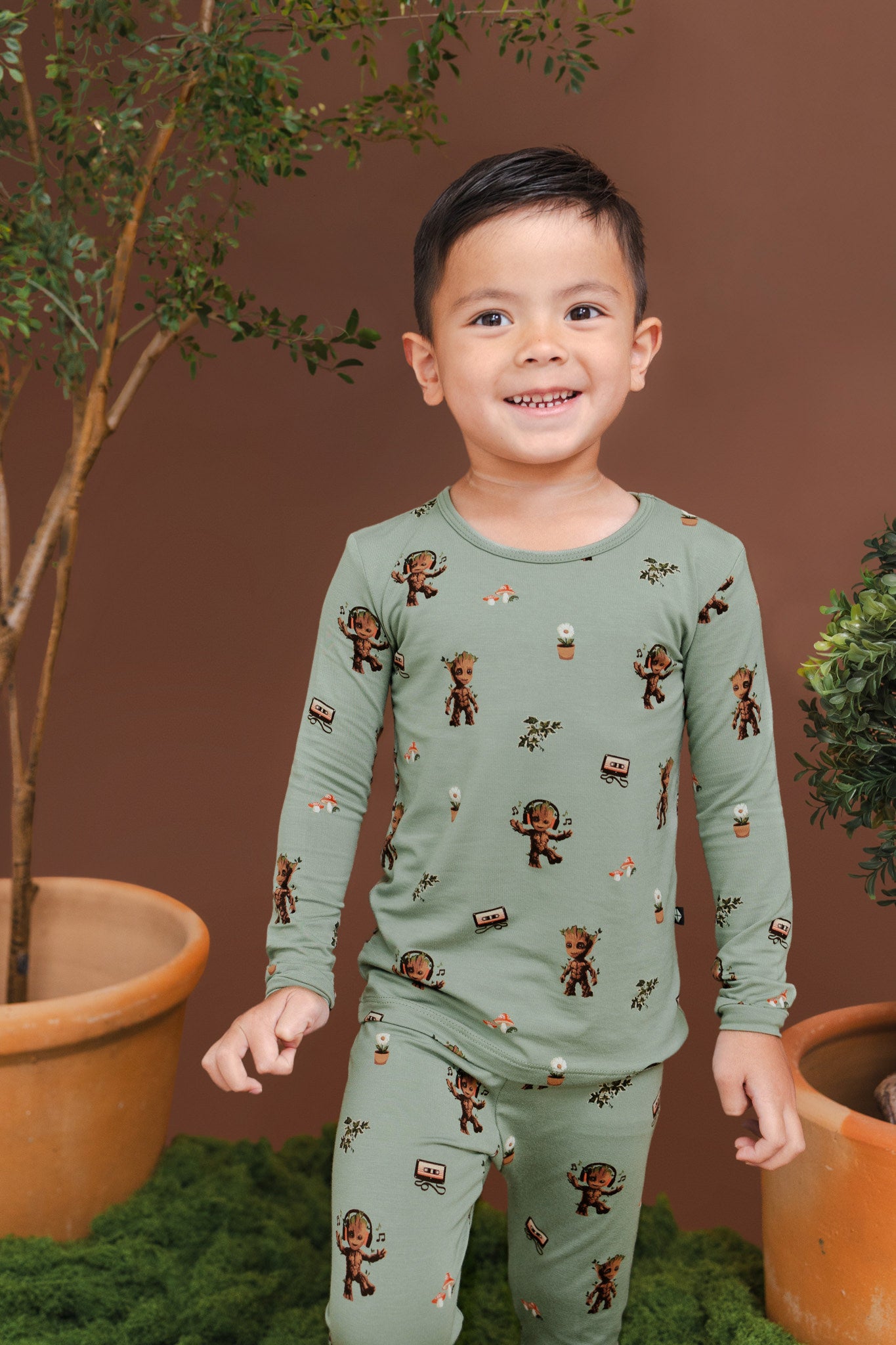 Boy smiling between planted pots wearing Green Long Sleeve Pajamas in Groot print