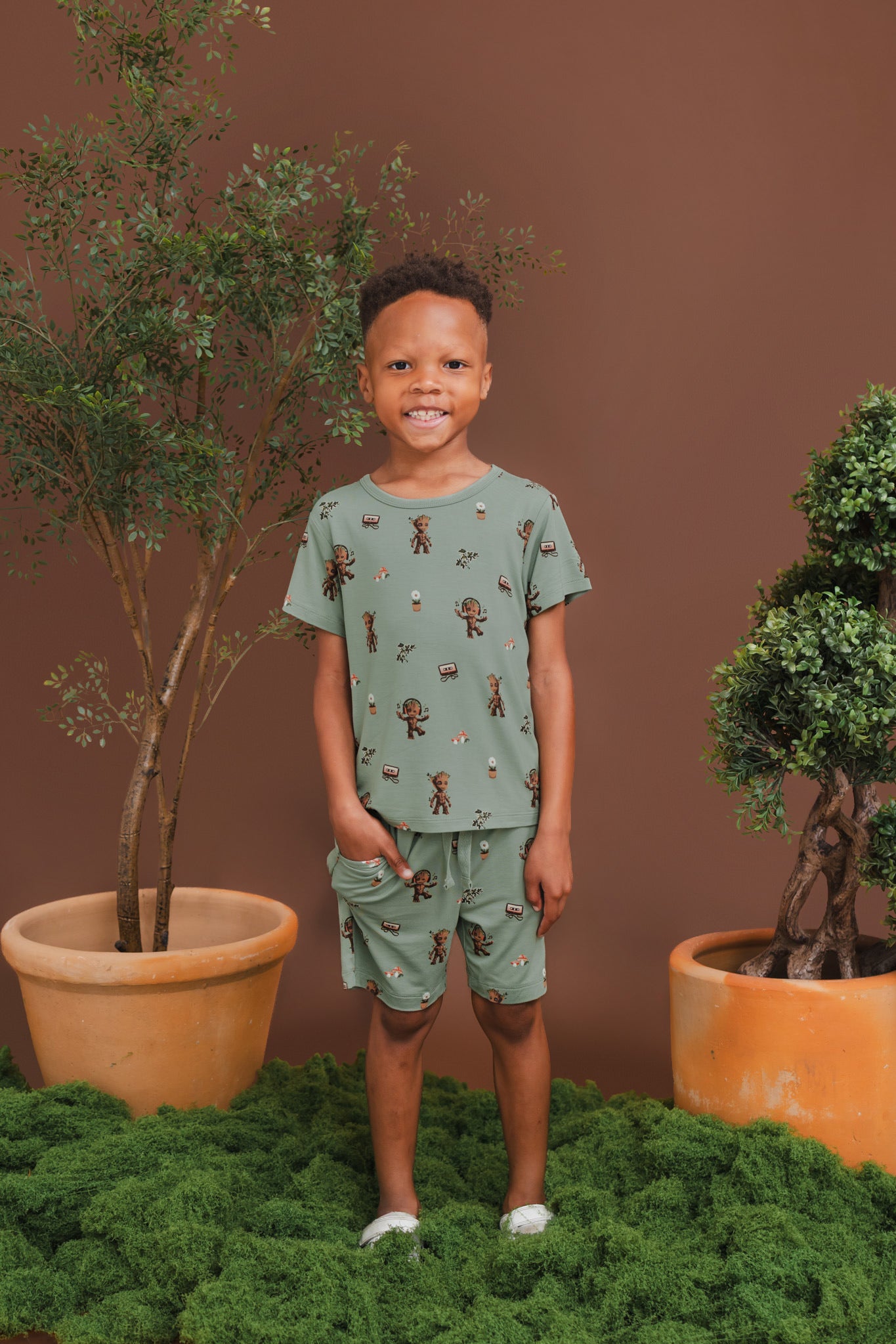 Boy wearing Green Toddler Basic Tee in Groot print over a similar print drawstring short standing on a grass and plant pots around