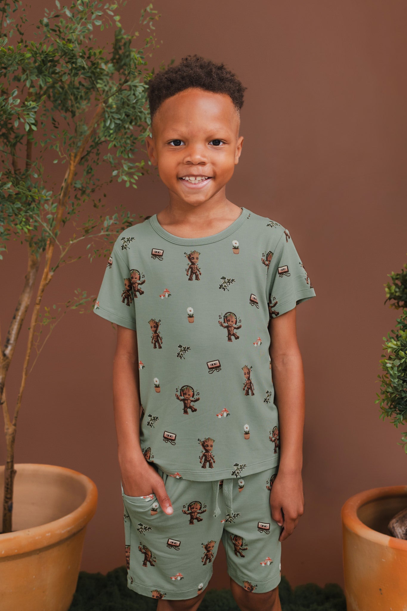 Boy wearing Green Toddler Basic Tee in Groot print over similar print drawstring shorts standing on a grass and plant pots around