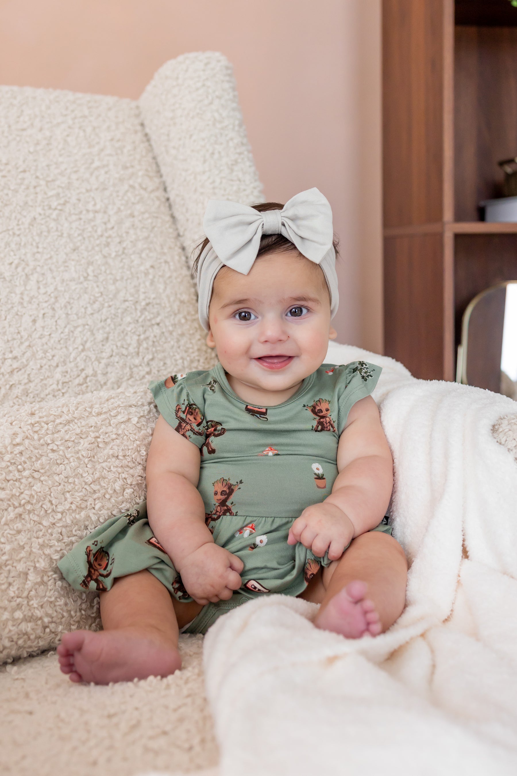 Baby sitting upright on a white sofa wearing Green Twirl Bodysuit Dress in Groot print and a beige knotted bow