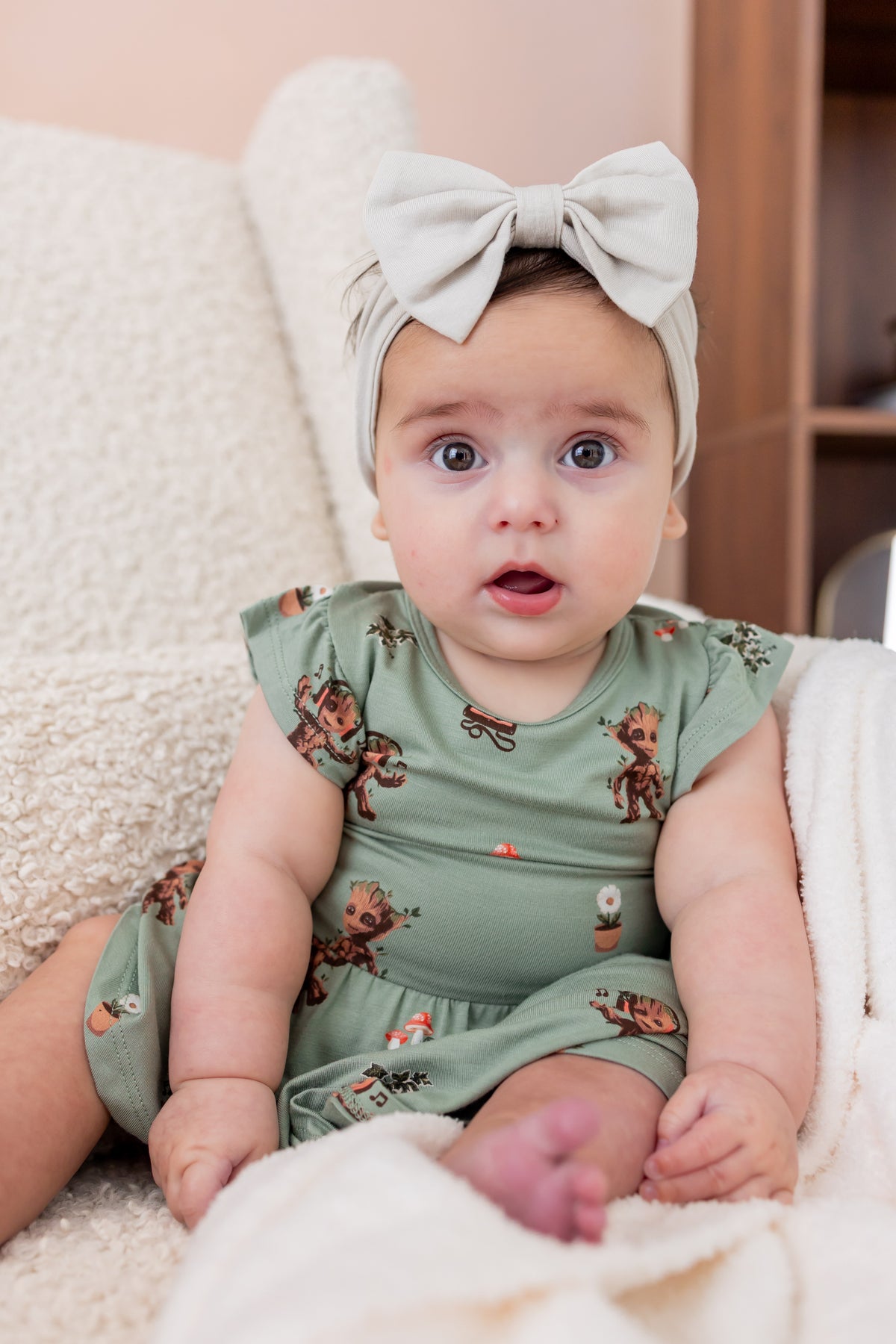 Baby sitting upright on a white sofa wearing Green Twirl Bodysuit Dress in Groot print and a beige knotted bow