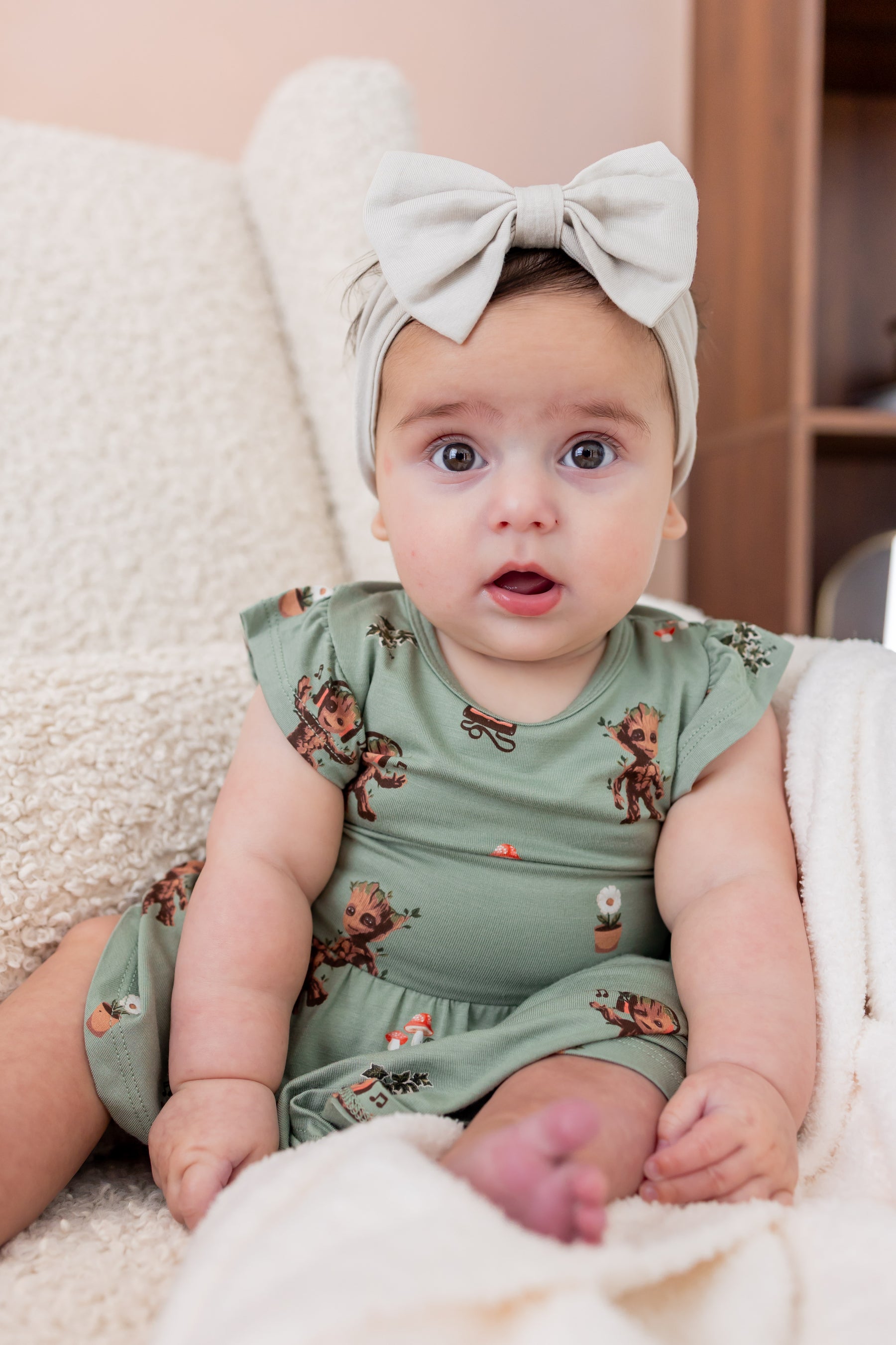 Baby sitting upright on a white sofa wearing Green Twirl Bodysuit Dress in Groot print and a beige knotted bow