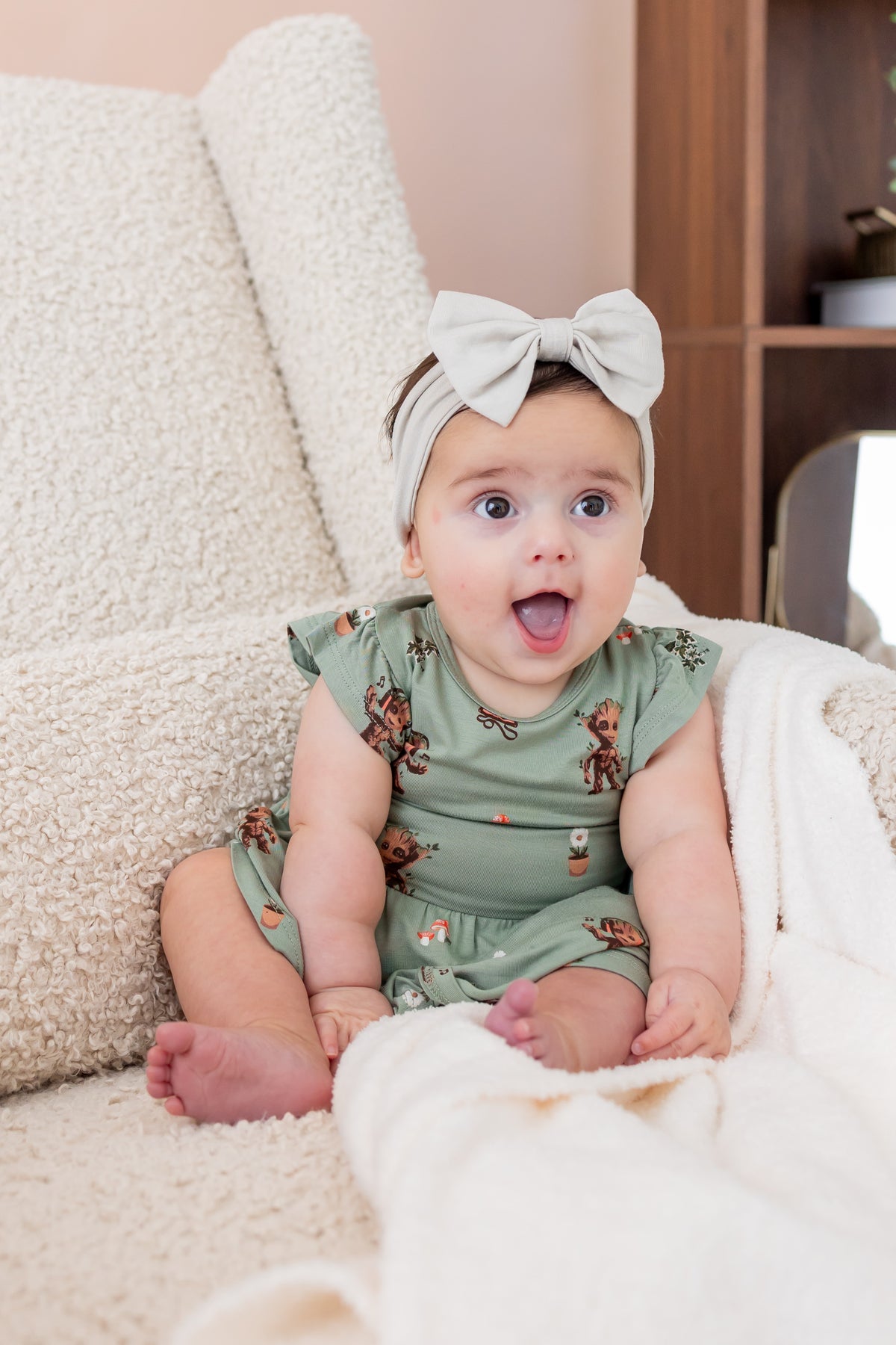 Baby smiling on a white sofa wearing Green Twirl Bodysuit Dress in Groot print and a beige knotted bow