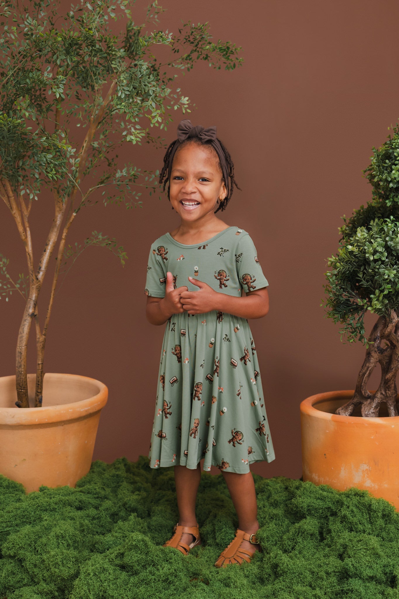 Girl wearing Green Twirl Dress in Groot print and brown bow standing between plant pots