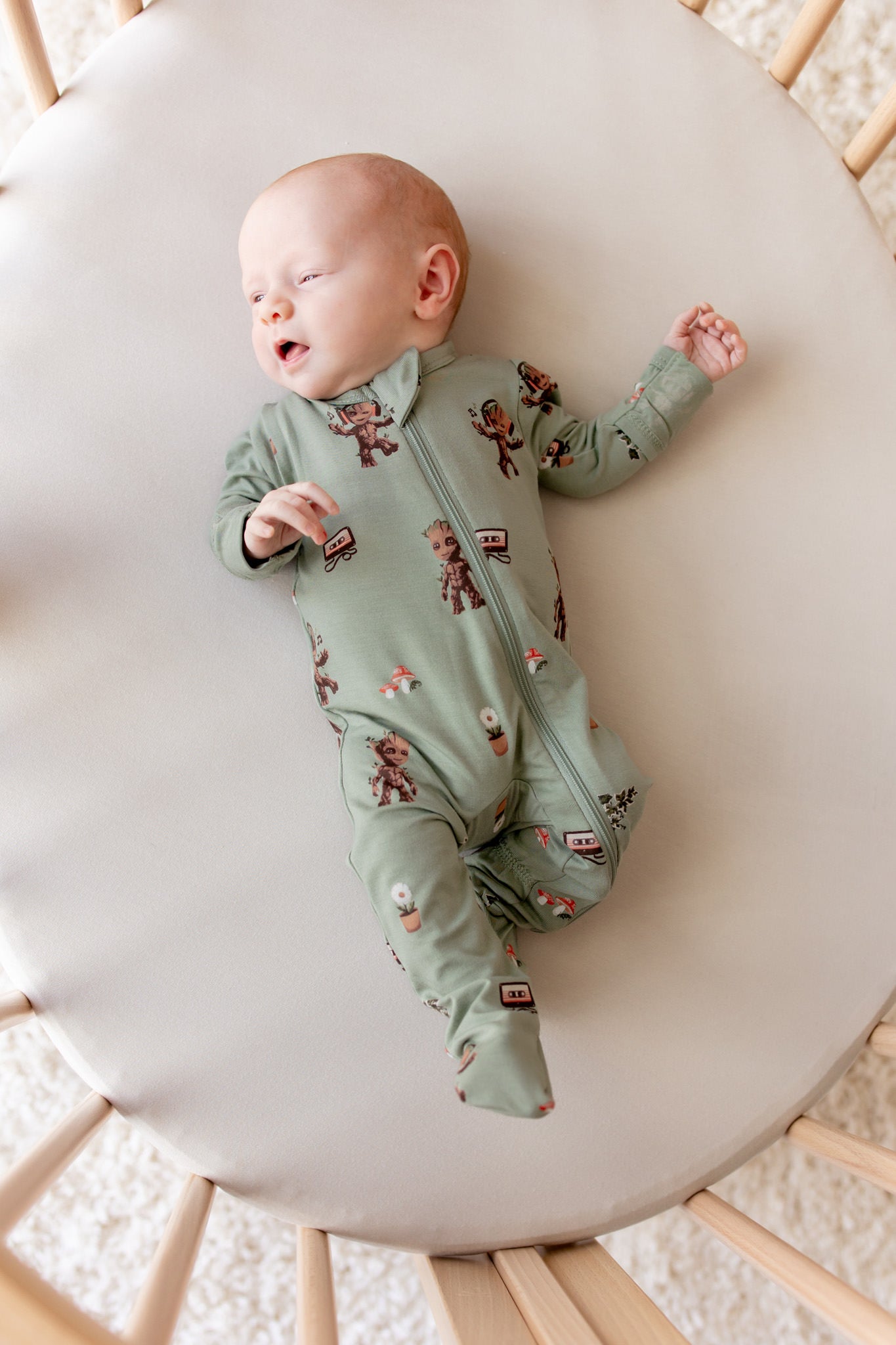 Baby sleeping in an oval Cot wearing Green Zippered Footie in Groot print on a beige bassinet sheet