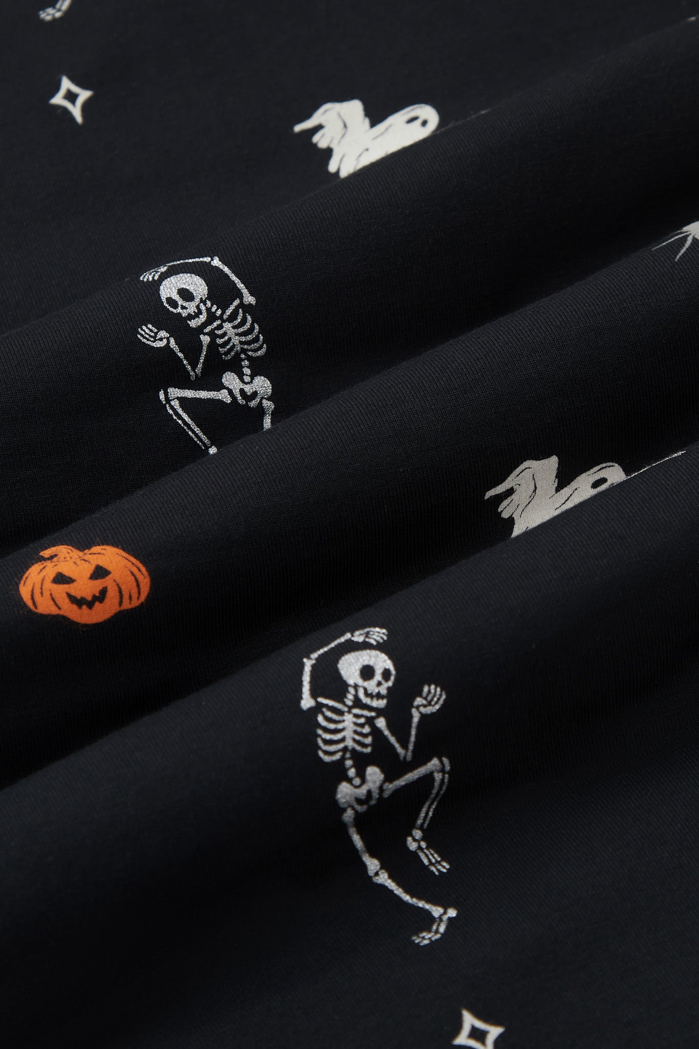 A repeating, all-over print featuring a flying white ghost, an orange jack o’ lantern, a dancing grey skeleton, a grey spider, and various white and grey twinkles, over a black background.