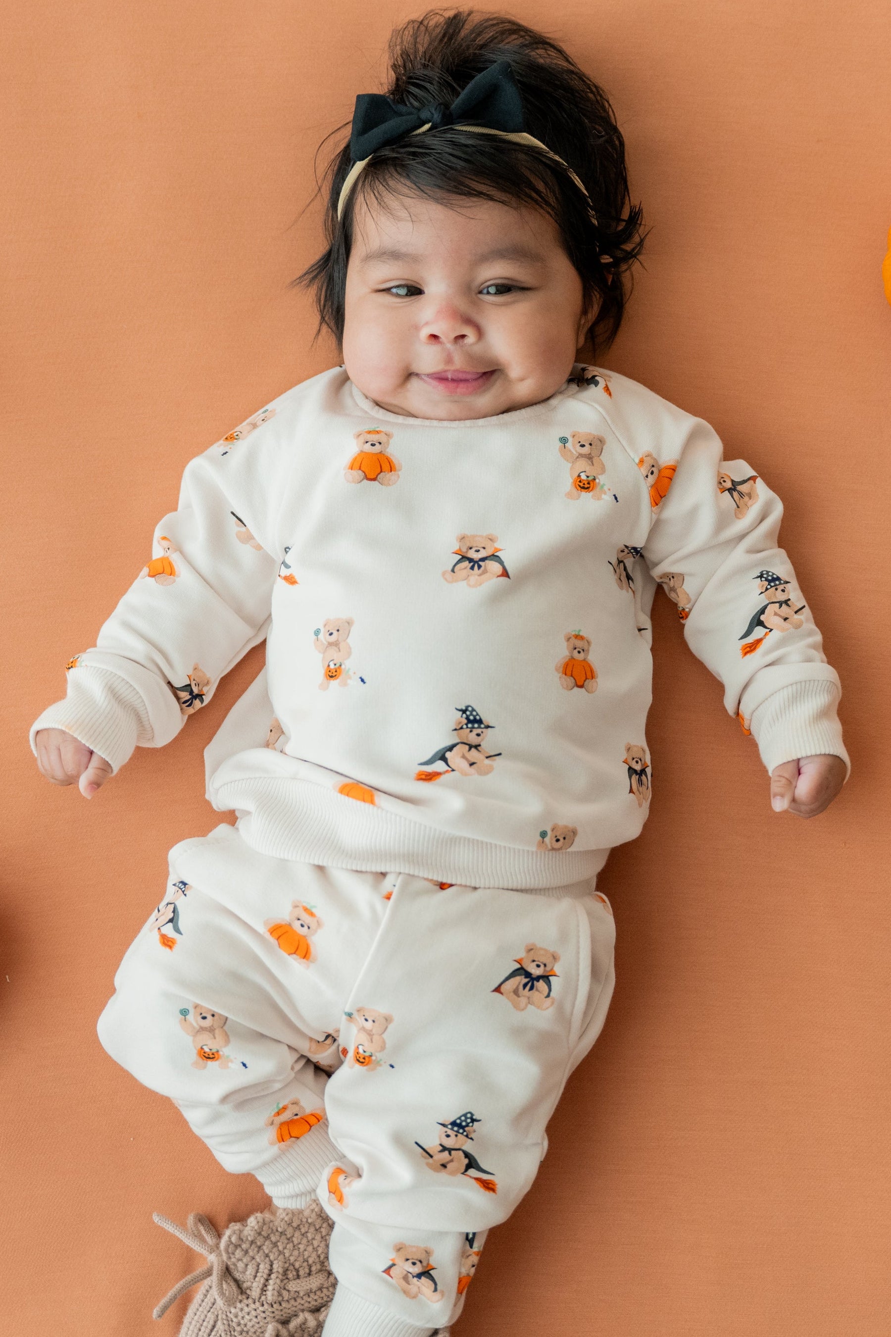 A baby lying on a peach background with small orange pumpkins around them, wearing an off-white long-sleeve pajama set with a Halloween print of ghosts, witches, and pumpkins. The baby is also wearing a black headband and knit socks.