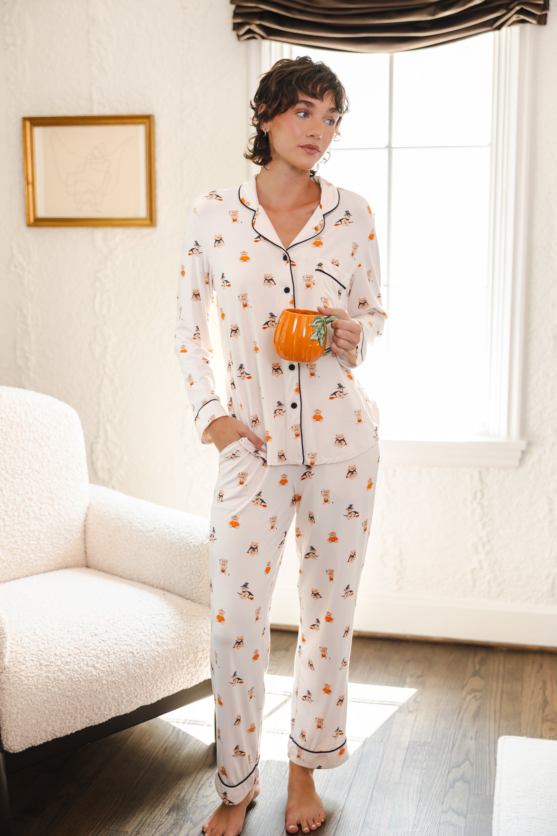 Modal wearing Beige Long-Sleeved Women's Pajama Set in Halloween Teddy print holding a mug standing in a living room