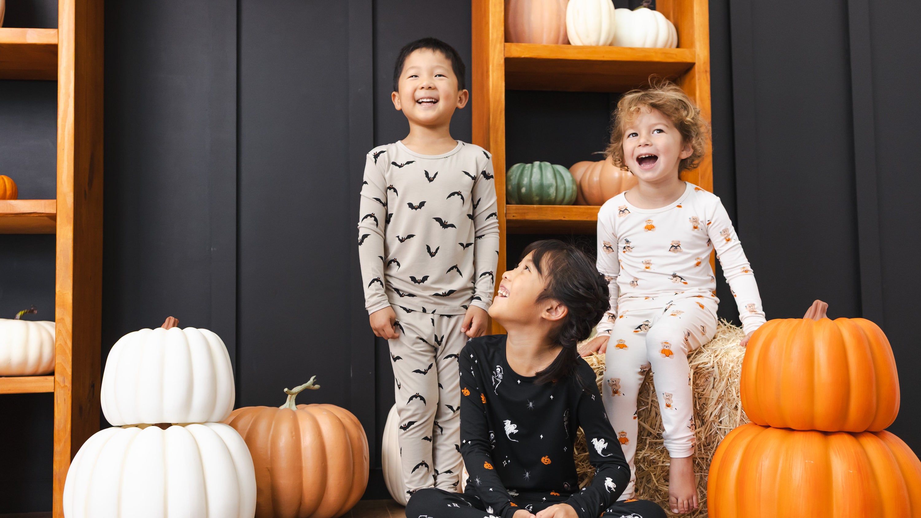 Three children wearing matching Halloween-themed pajamas pose together in front of a dark paneled wall with wooden shelves and decorative pumpkins. The children's pajamas feature spooky prints: one light gray with black bats, one white with halloween teddy bears, and one black with white ghosts, skeletons and pumpkins.