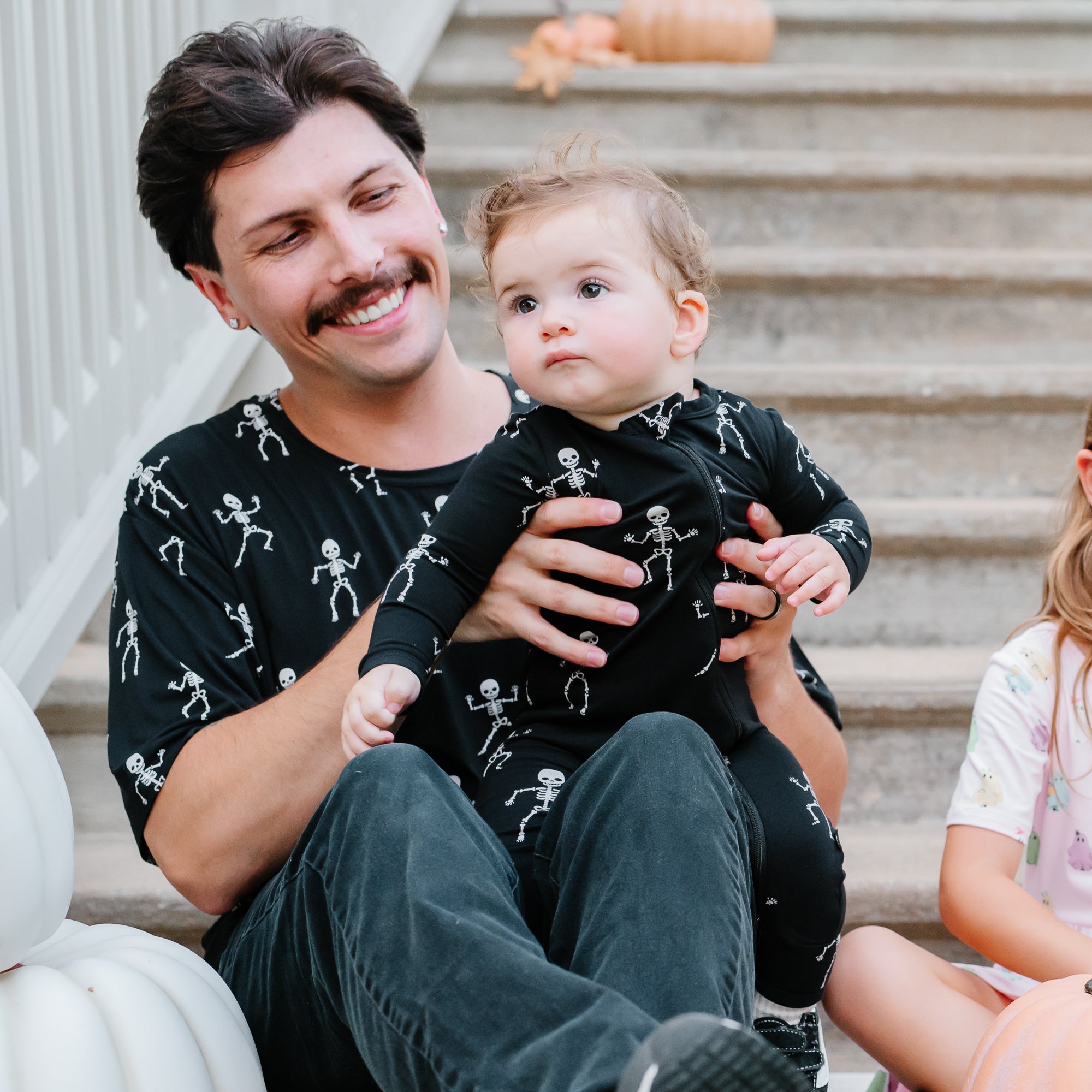 Man holding a baby in skeleton-themed outfits on a staircase with pumpkins.