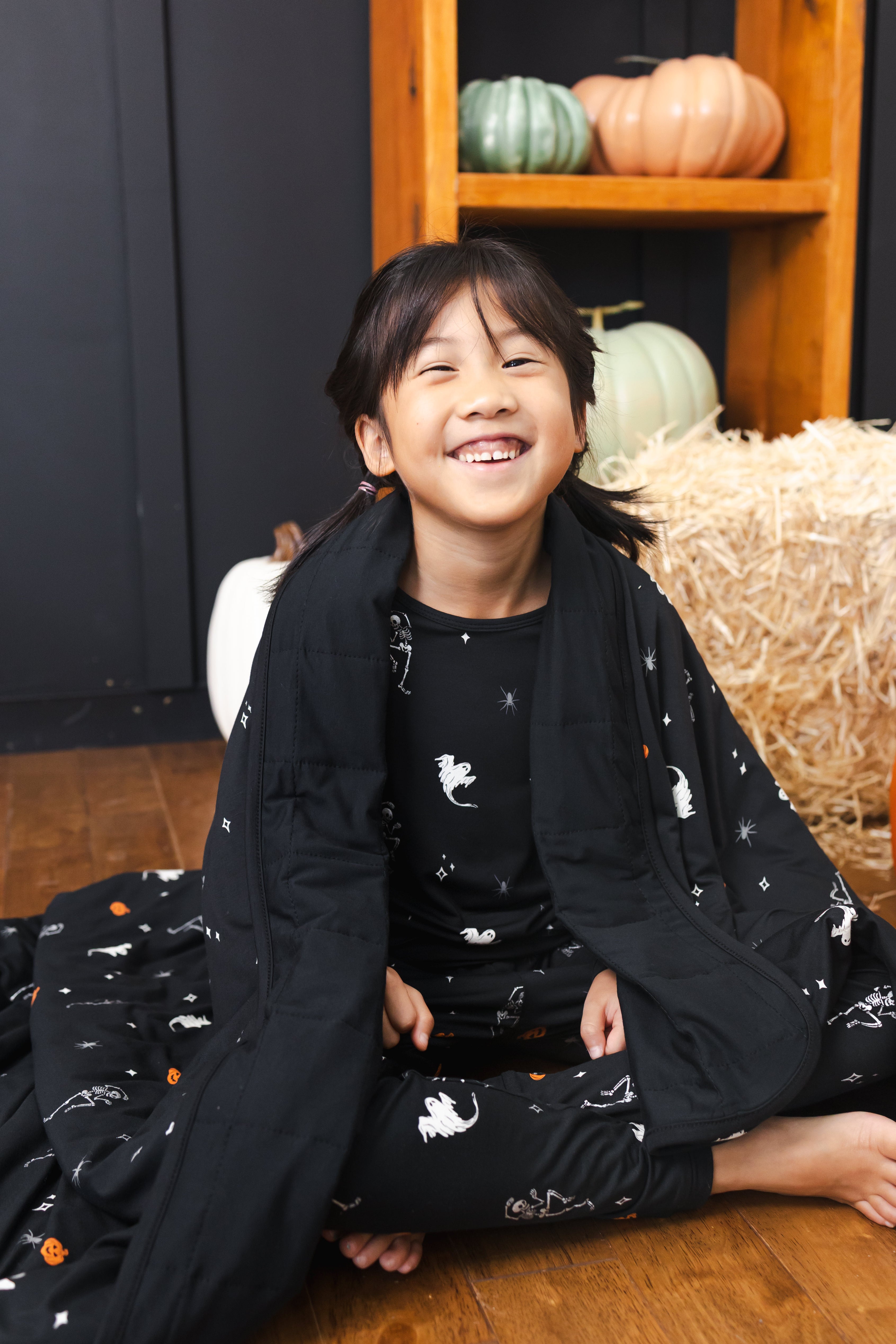 Girl wrapped in Toddler Blanket in Halloween Friends 1.0 print wearing a similar print long sleeve pajama set sitting in front of Halloween decorations