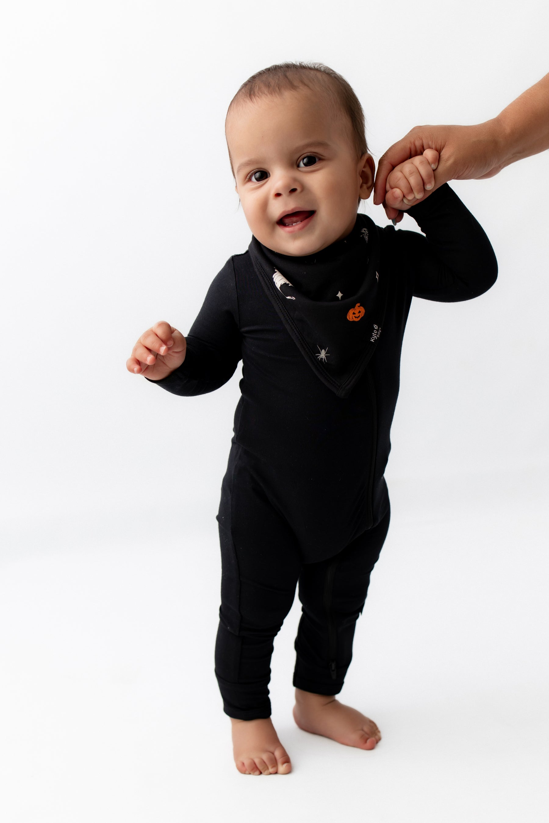 Baby wearing Bib in Halloween Friends print over a black romper holding an adult's hand on a white background