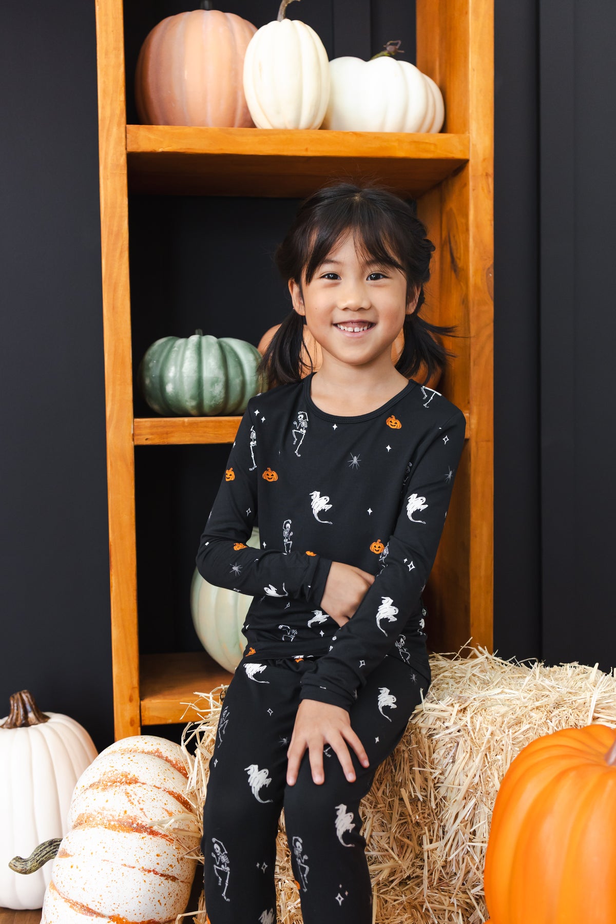 Girl smiling in black Long Sleeve Pajamas in Halloween Friends print sitting with pumpkin decorations