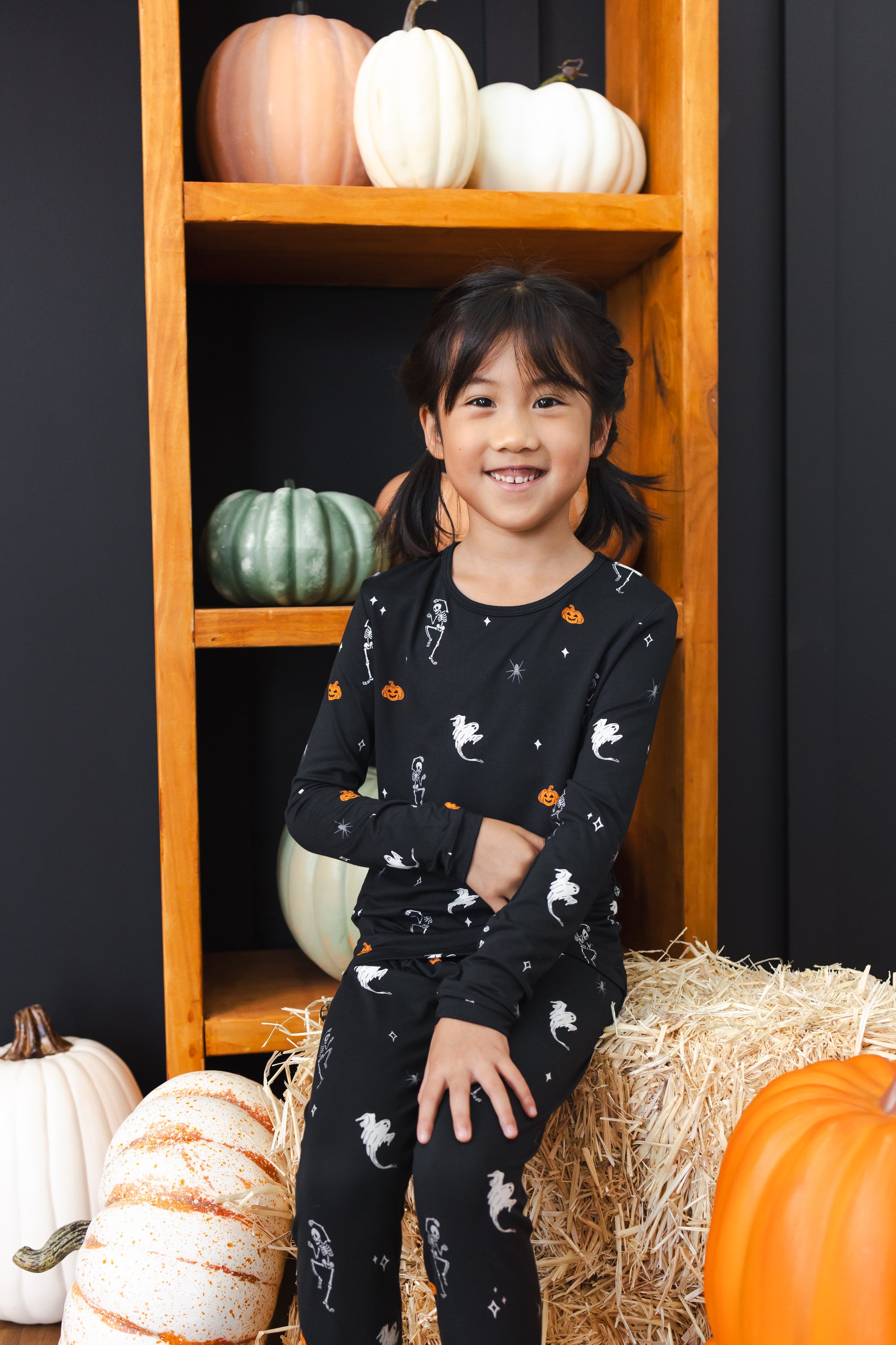 Girl smiling in black Long Sleeve Pajamas in Halloween Friends print sitting with pumpkin decorations