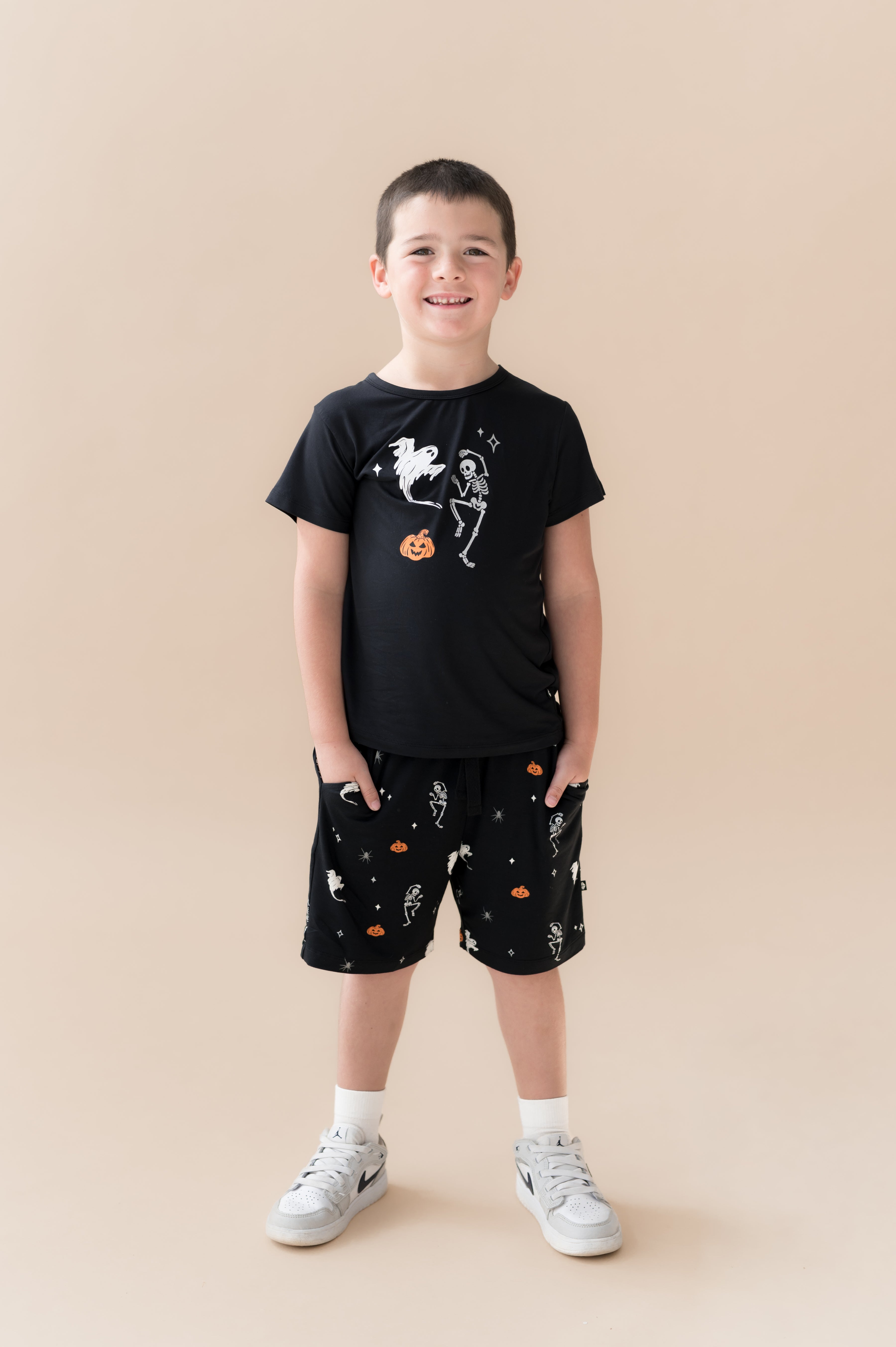 Boy wearing Black Toddler Basic Tee in Halloween Friends print in center over Similar print Drawstring shorts and white shoes on a beige background
