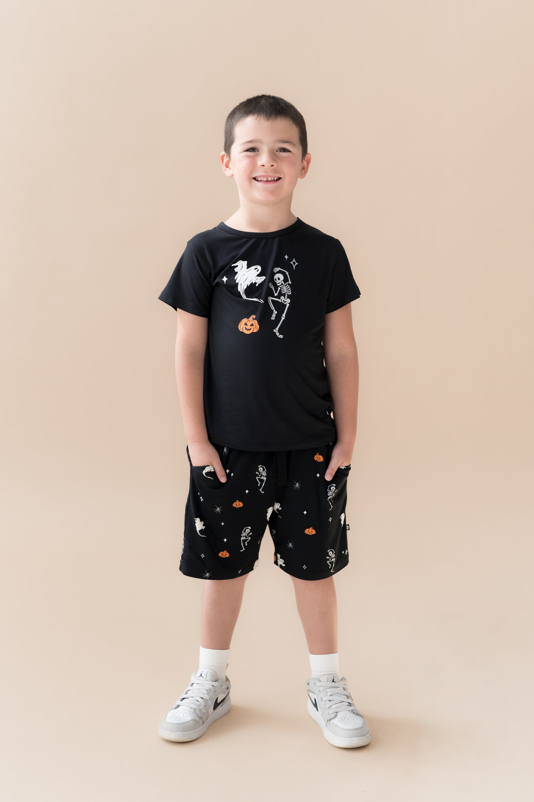 Boy wearing Black Toddler Basic Tee in Halloween Friends print in center over Similar print Drawstring shorts and white shoes on a beige background