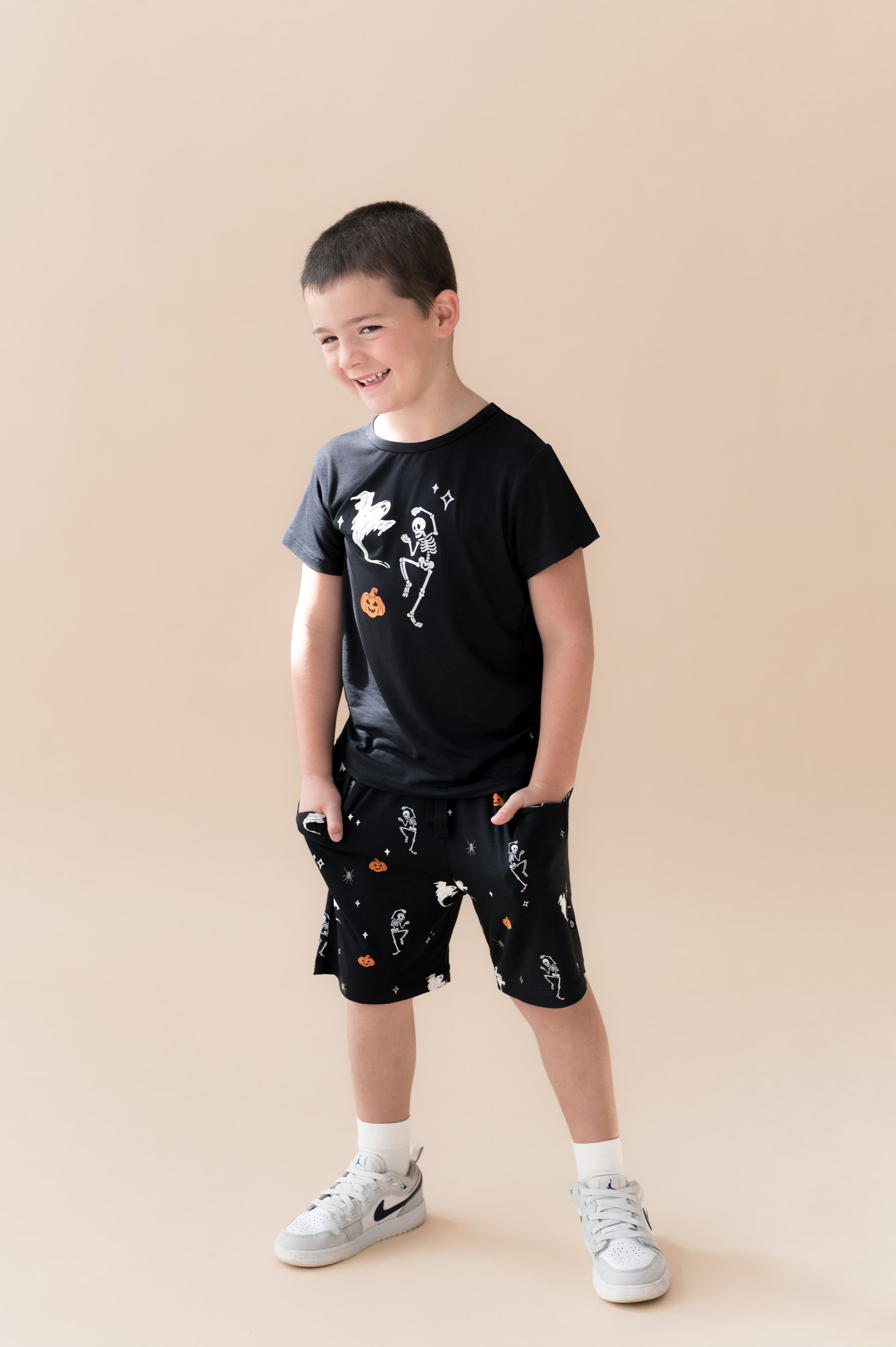 Boy posing in Black Toddler Basic Tee in Halloween Friends print in center over Similar print Drawstring shorts and white shoes on a beige background