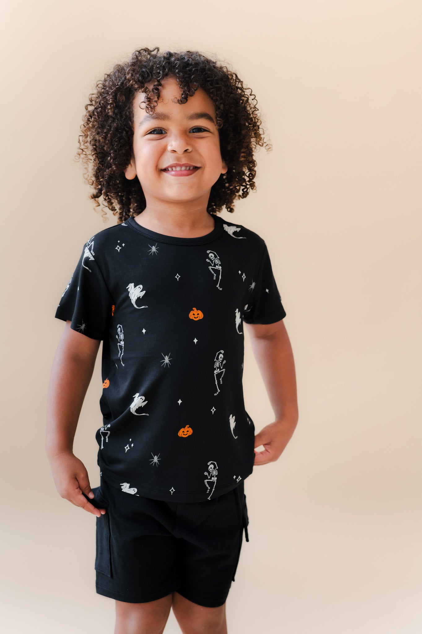 Boy smiling in Toddler Basic Tee in Halloween Friends print over black shorts on a beige background