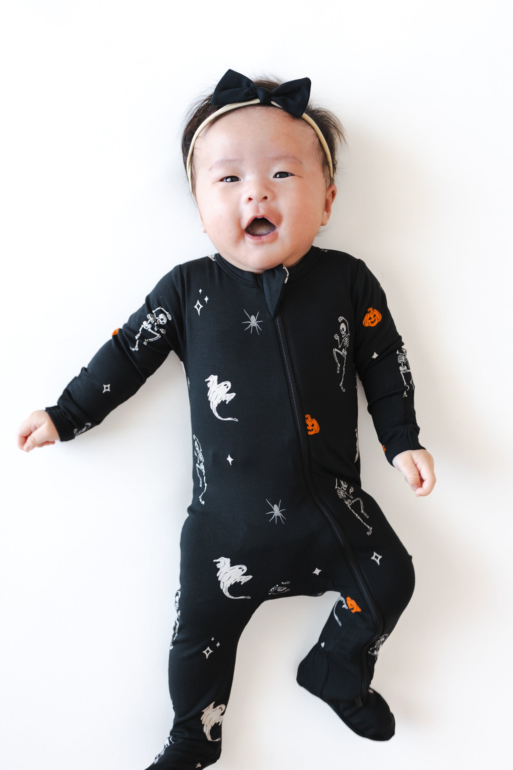 Baby wearing Zippered Footie in Halloween Friends print and a black bow on a white background
