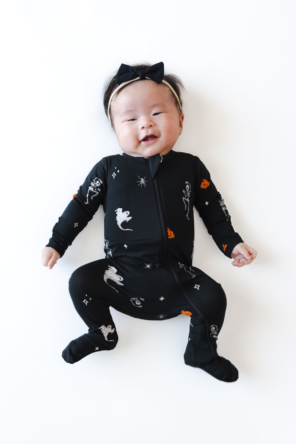 Baby wearing Zippered Footie in Halloween Friends print and a black bow on a white background