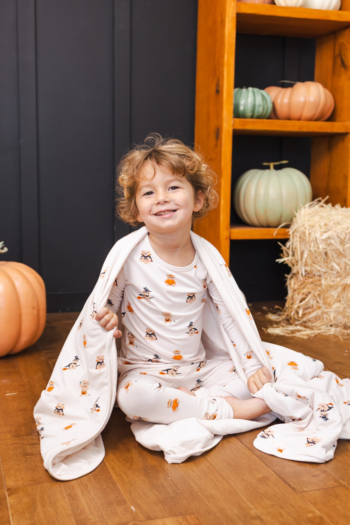 Boy wrapped in a Toddler Blanket in Halloween Teddy 1.0 print wearing a similar print long sleeve pajama set sitting in between the pumpkin decorations 