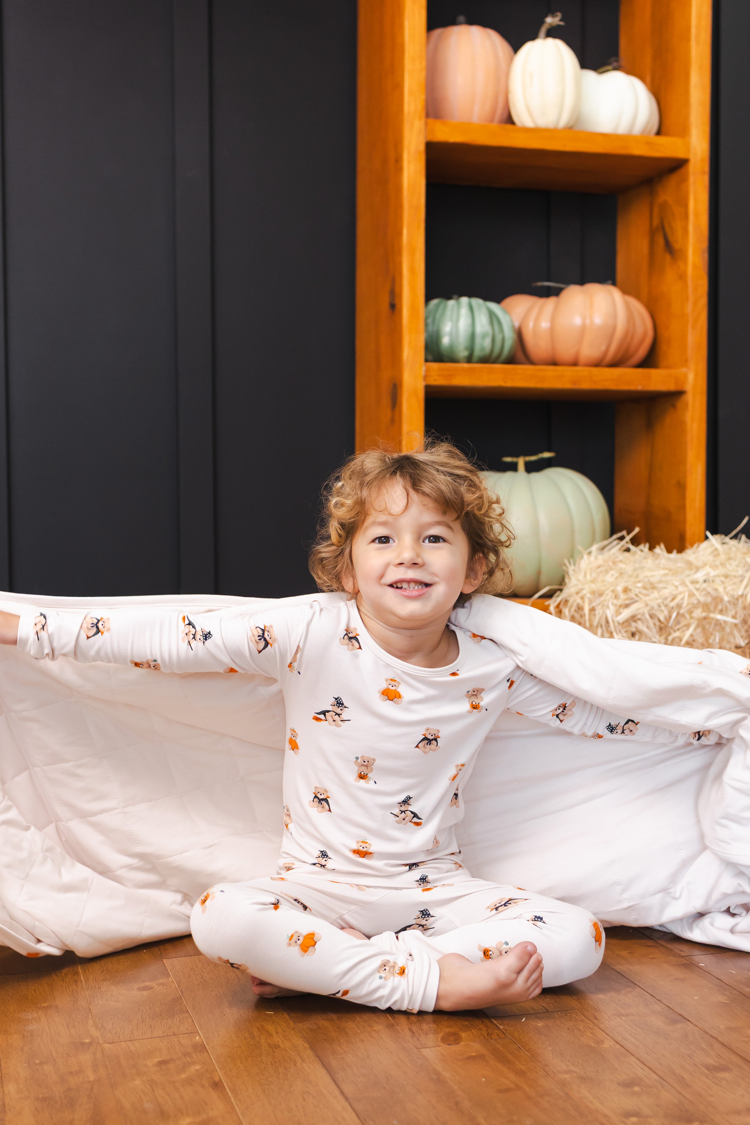 Boy holding Toddler Blanket in Halloween Teddy 1.0 print wearing a similar print long sleeve pajama set with pumpkin decoration in the background