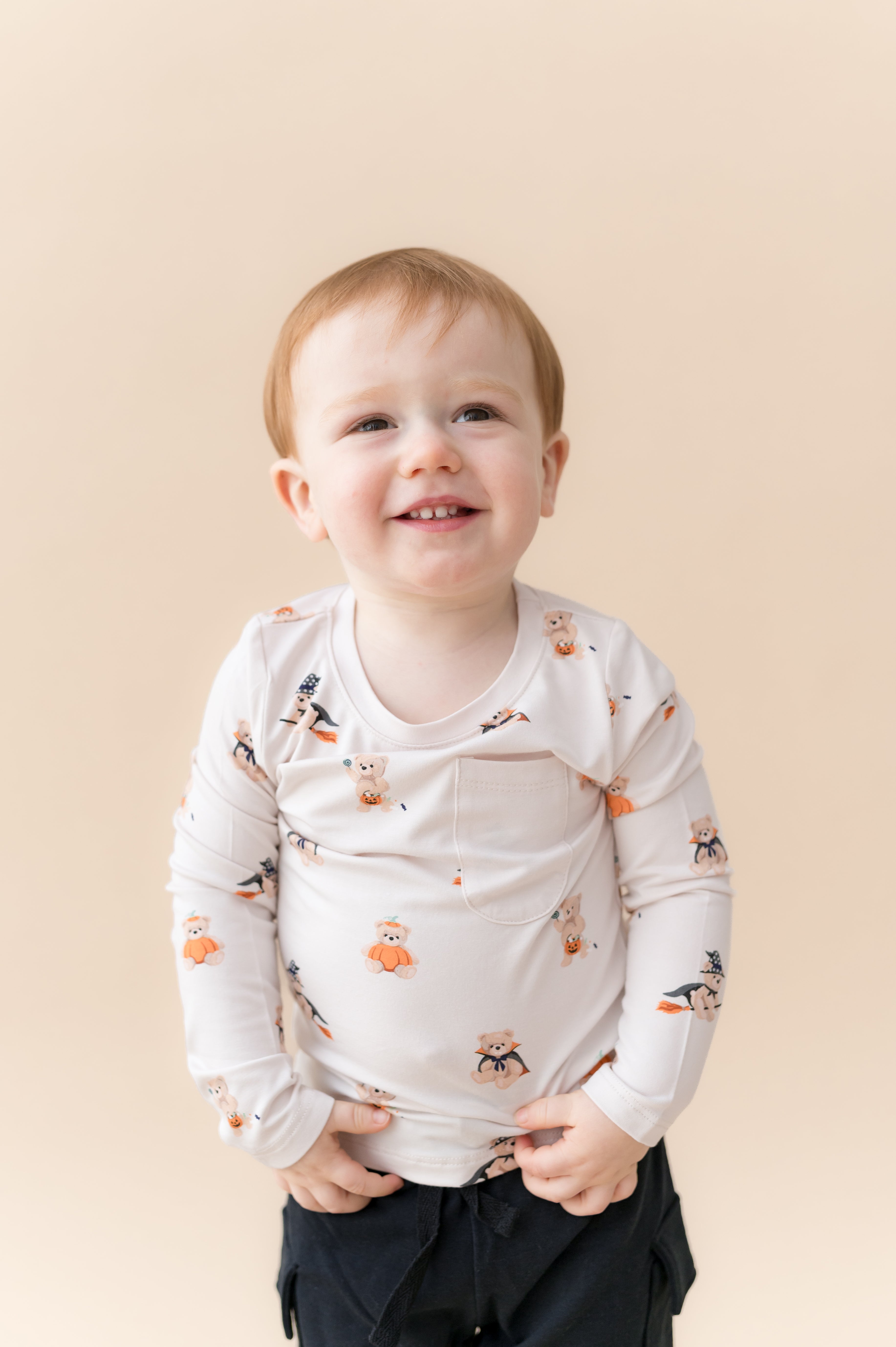 Boy wearing Long Sleeve Toddler Crew Neck Tee in Halloween Teddy print over black shorts on a beige background