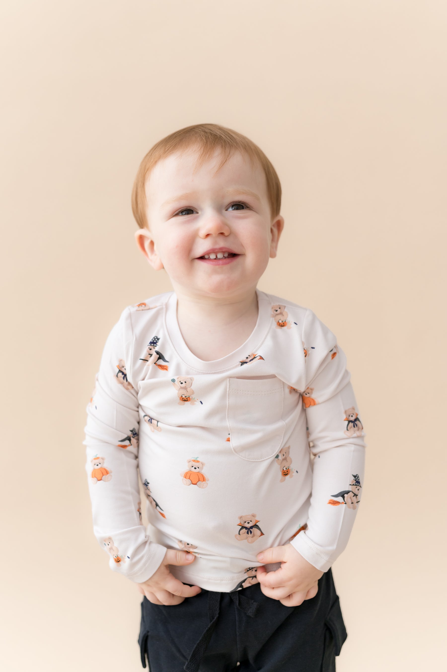 Boy wearing Long Sleeve Toddler Crew Neck Tee in Halloween Teddy print over black shorts on a beige background