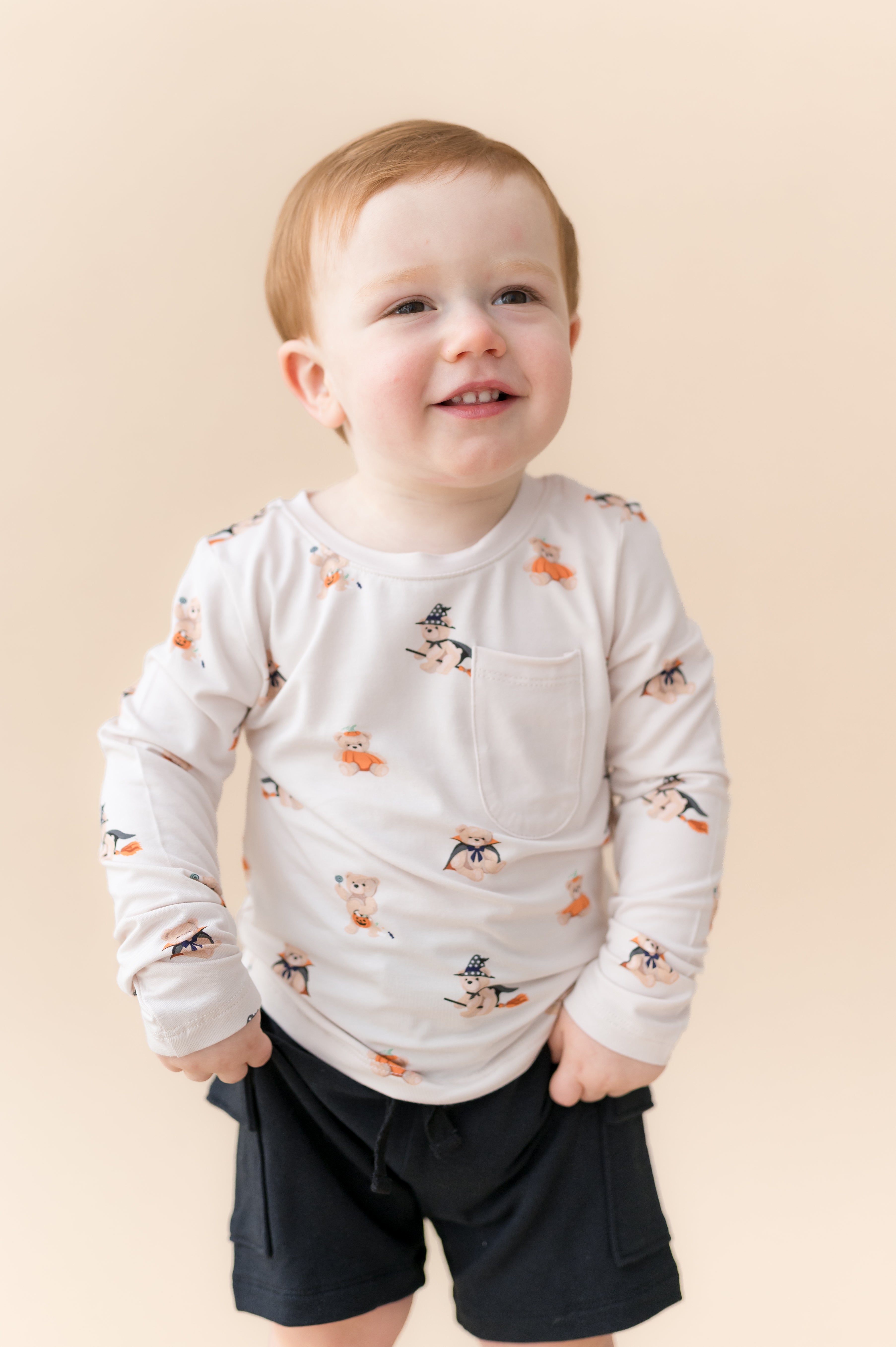 Boy posing in Long Sleeve Toddler Crew Neck Tee in Halloween Teddy print over black shorts on a beige background
