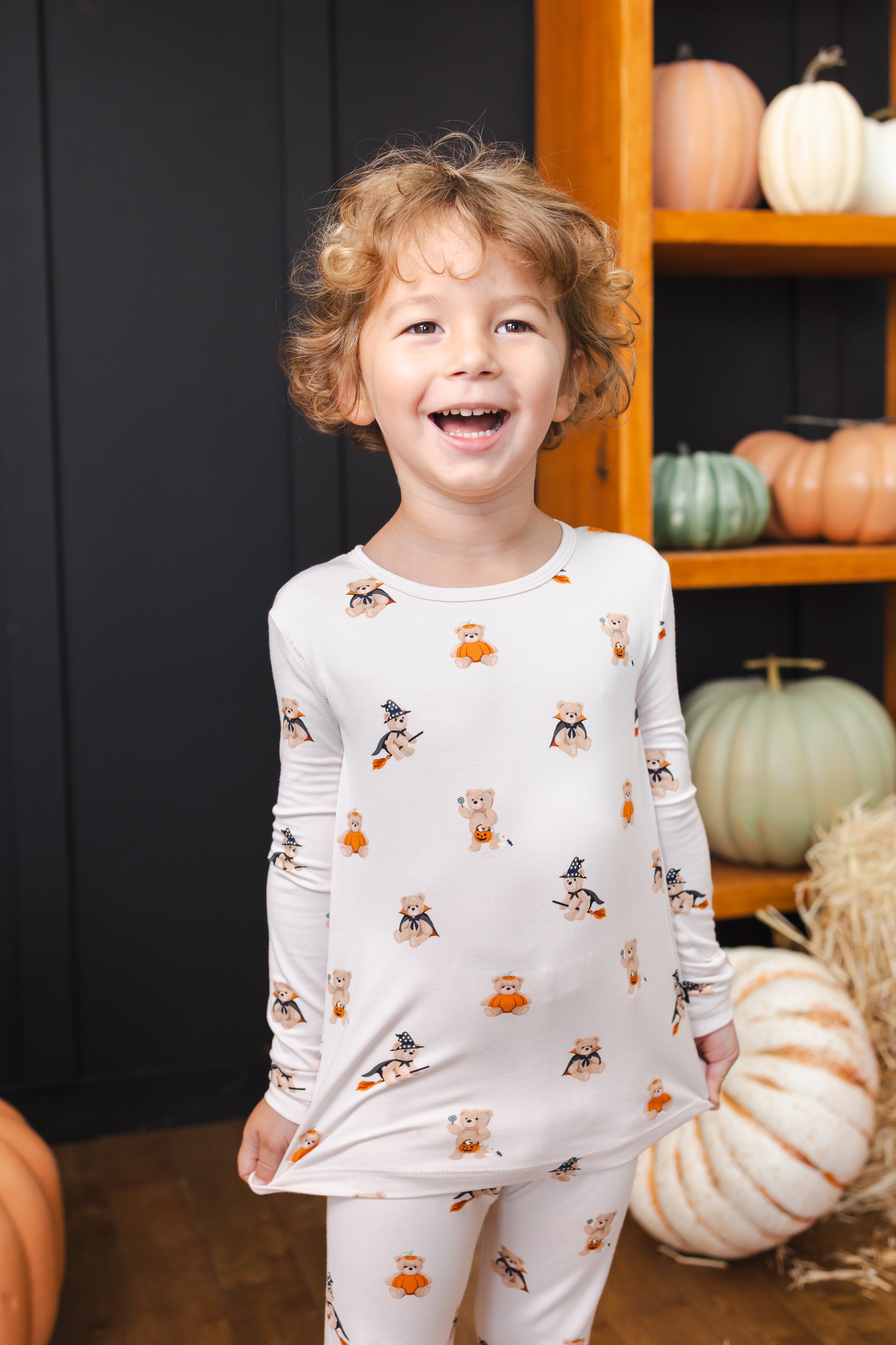 Boy laughing in Long Sleeve Pajamas in Halloween Teddy print standing in front of Halloween pumpkin decorations
