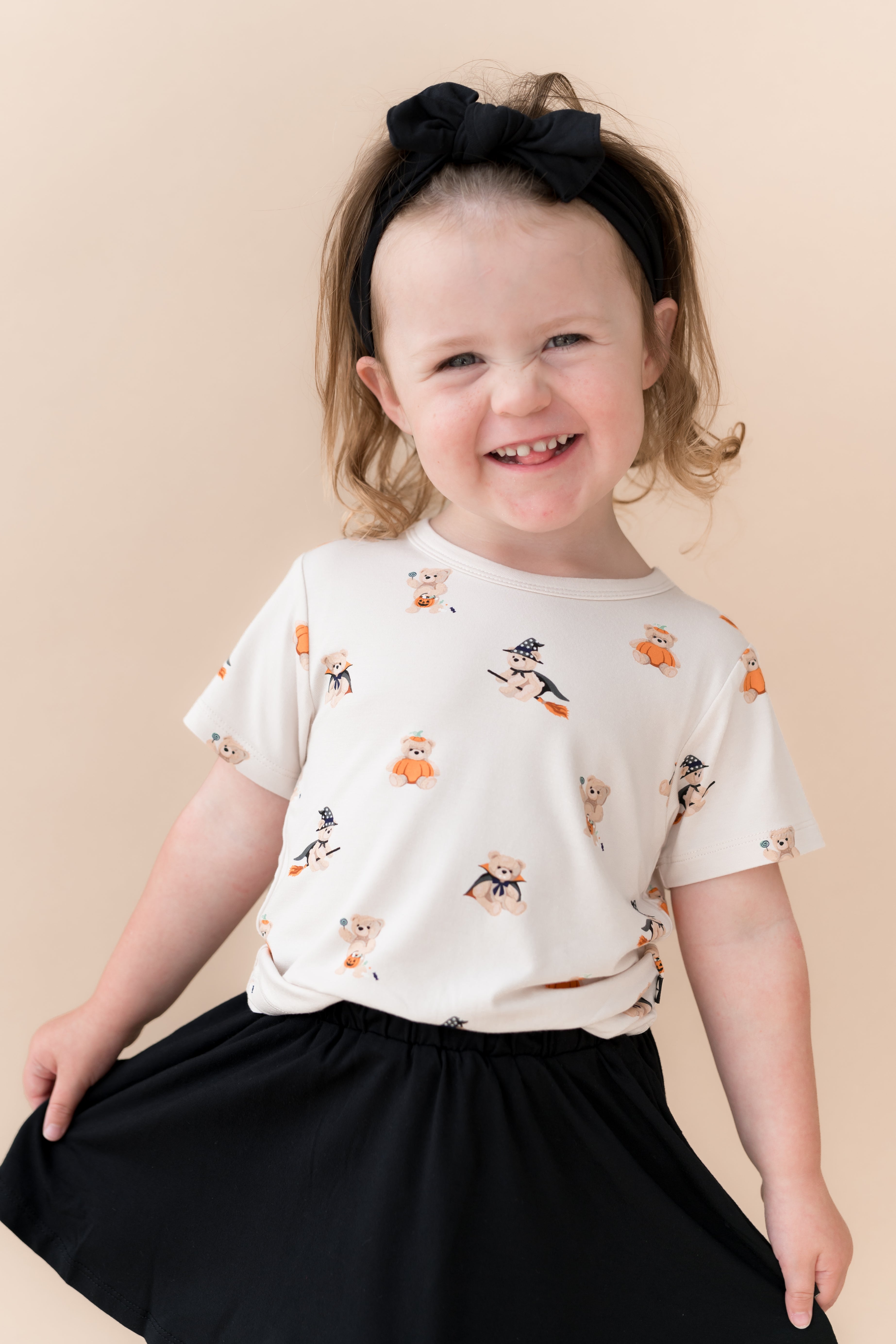 Girl smiling in Toddler Basic Tee in Halloween Teddy print over Black skort shoes on a beige background