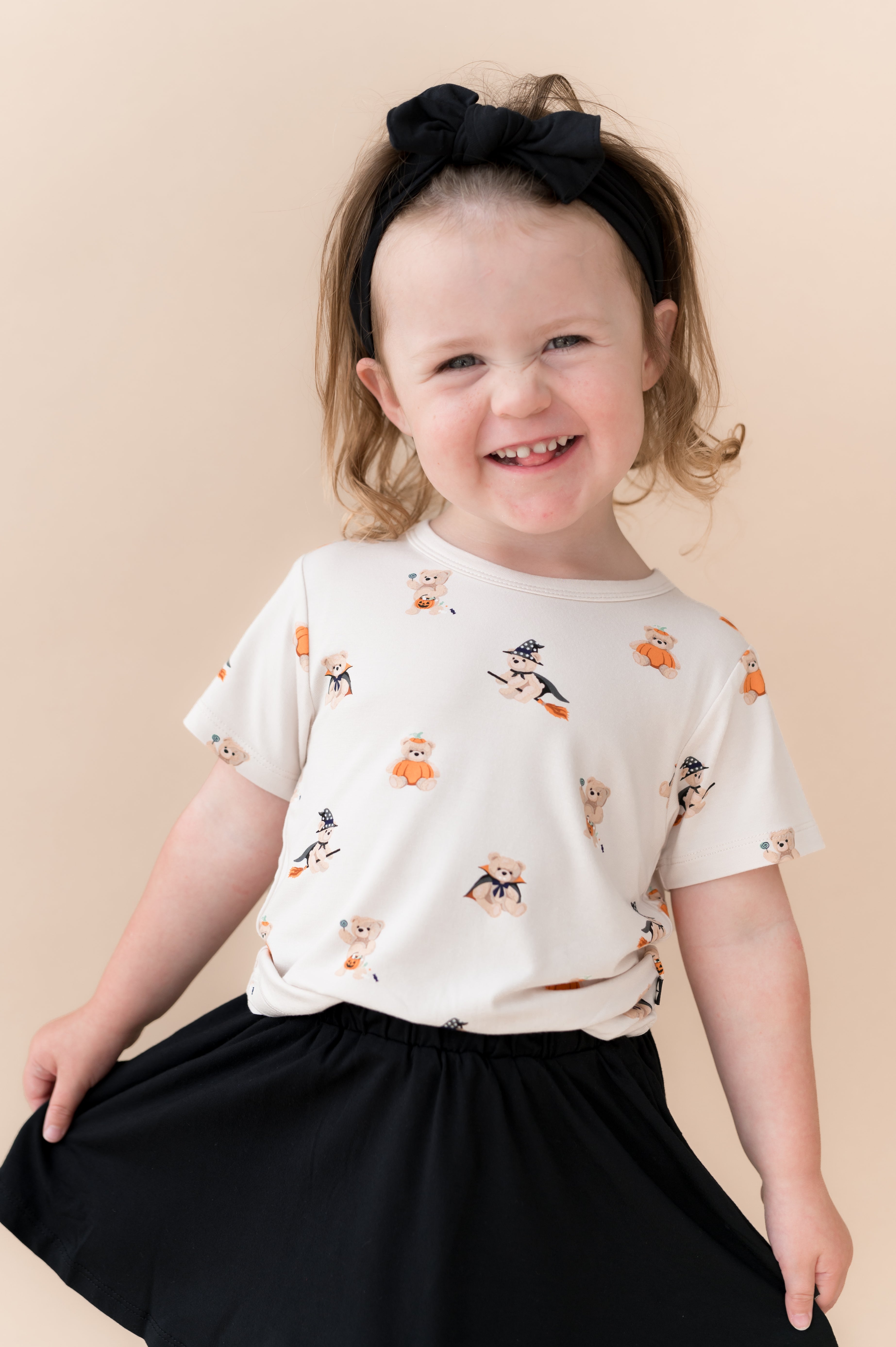 Girl smiling in Toddler Basic Tee in Halloween Teddy print over Black skort shoes on a beige background