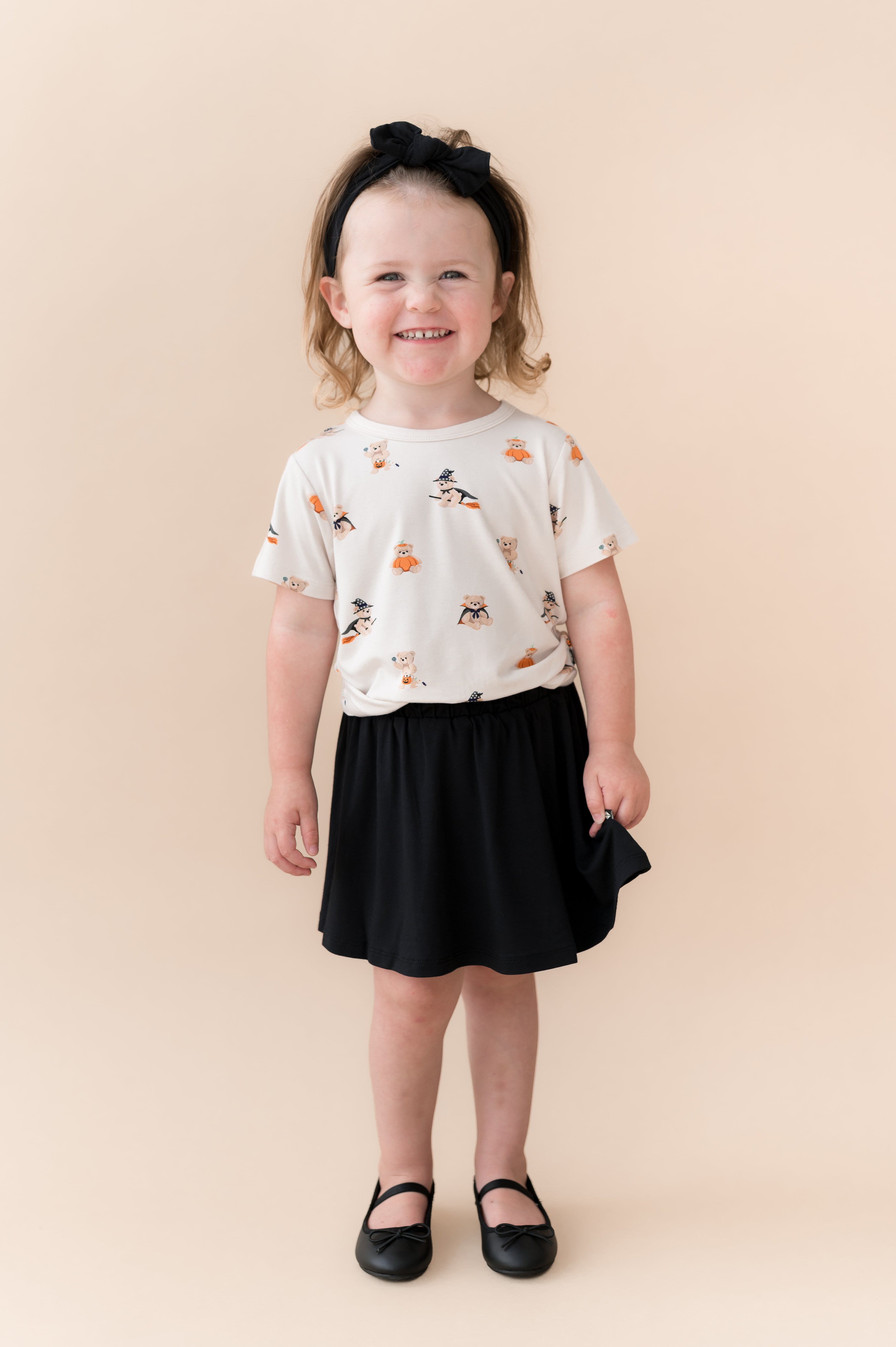 Girl wearing Toddler Basic Tee in Halloween Teddy print over Black skort shoes and bow on a beige background