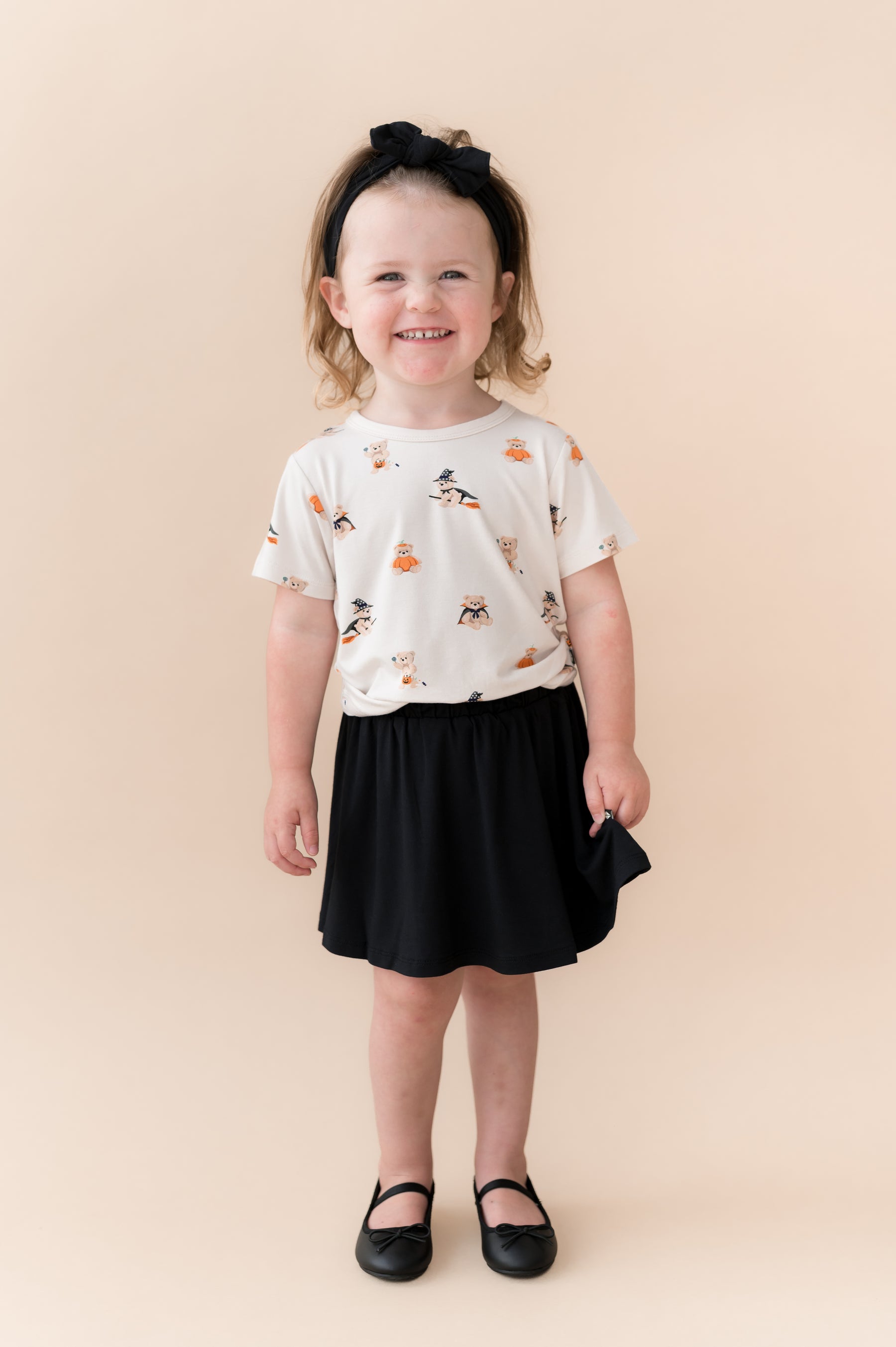 Girl wearing Toddler Basic Tee in Halloween Teddy print over Black skort shoes and bow on a beige background
