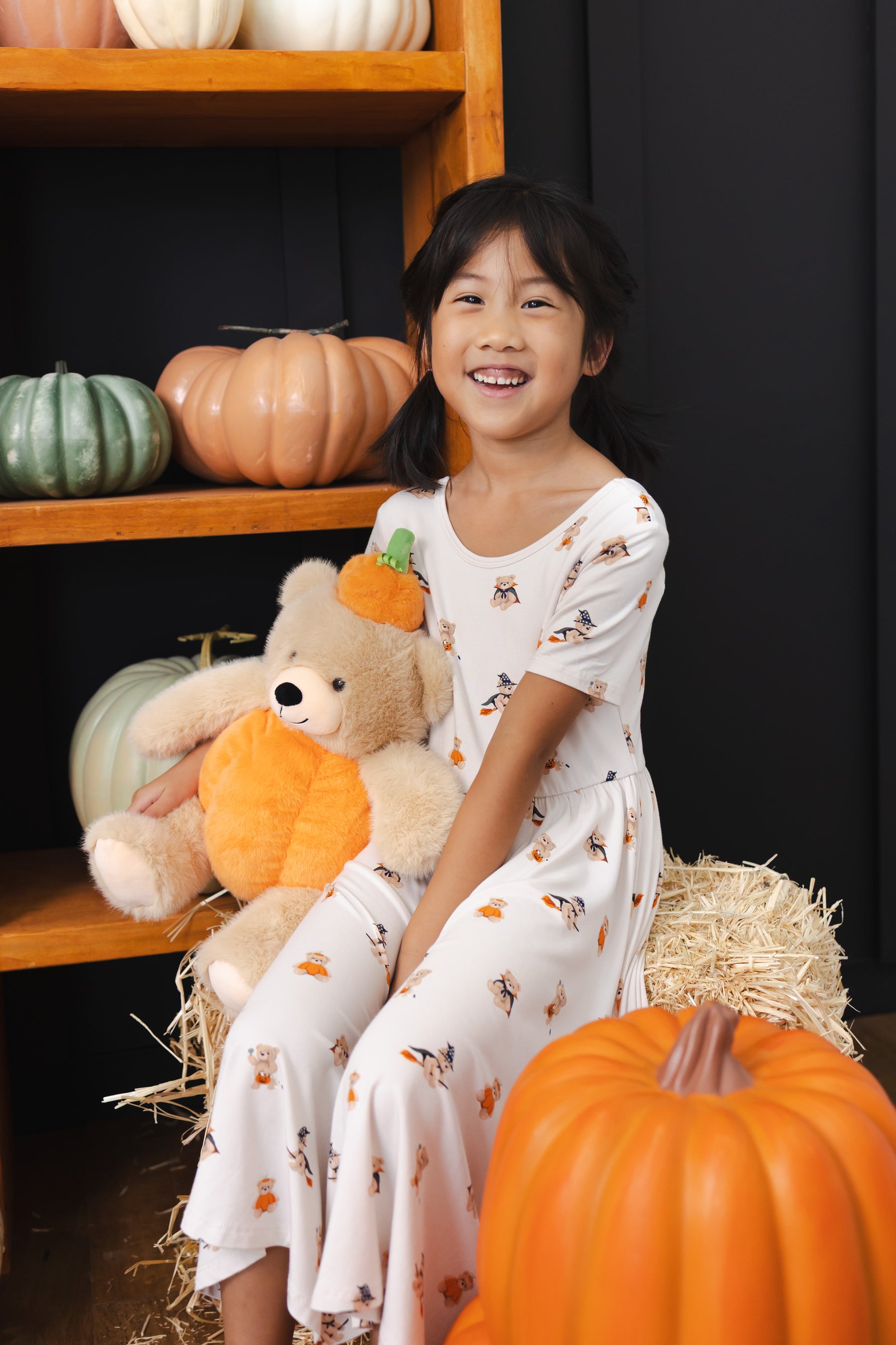 Girl sitting with a stuffed teddy bear wearing Twirl Dress in Halloween Teddy print with pumpkin decorations around